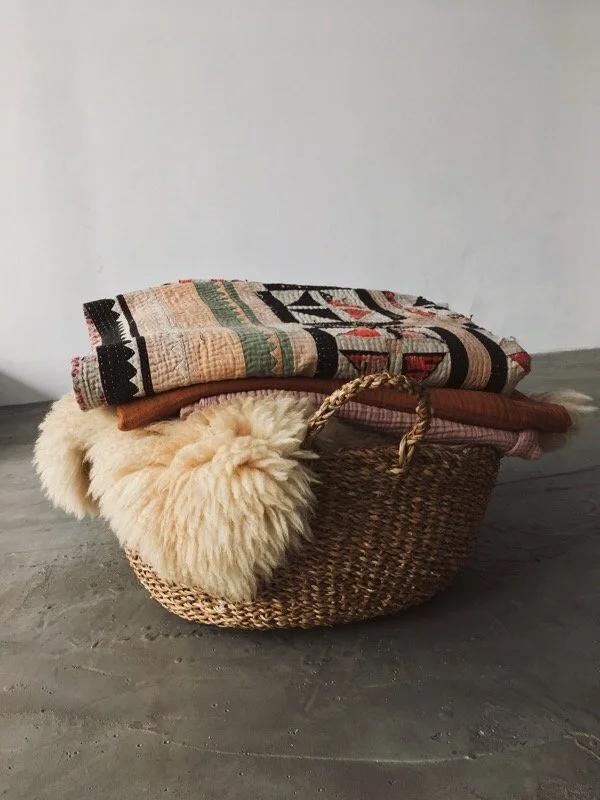 A woven basket filled with folded blankets and a sheepskin throw, placed on a wooden floor against a plain wall.