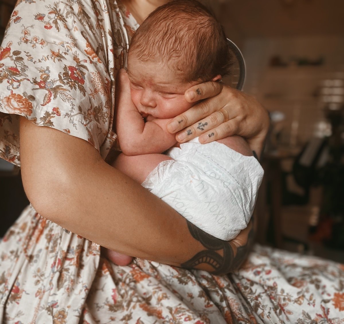 A person holding a newborn baby close, with the baby's face resting against their hand. The person has tattoos on their fingers and arm, and is wearing a floral dress. The baby has a fair complexion and reddish hair.