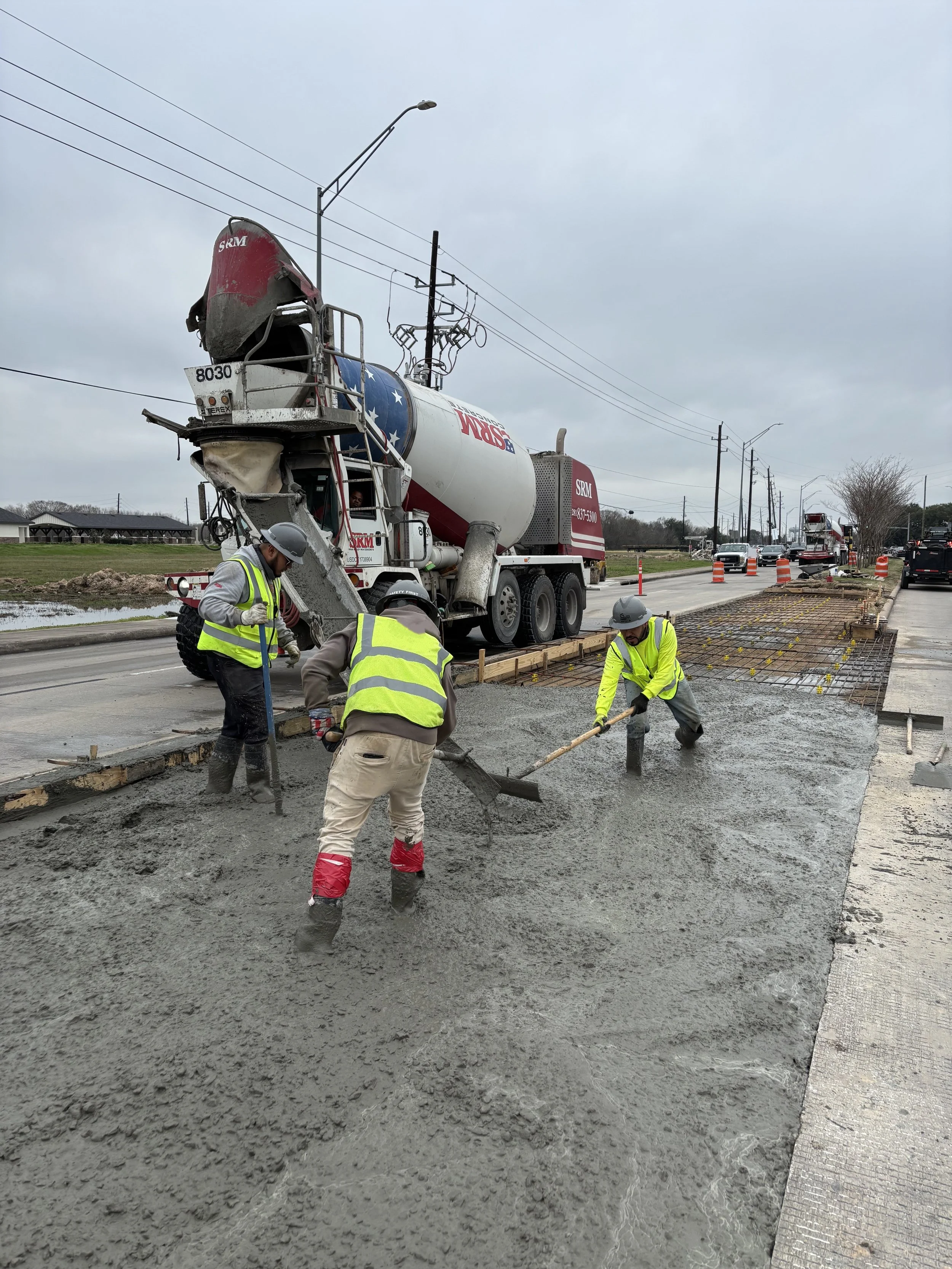 Construction workers pouring and leveling wet concrete for a sidewalk or road extension, with a concrete mixer truck in the background and orange traffic cones along the street.