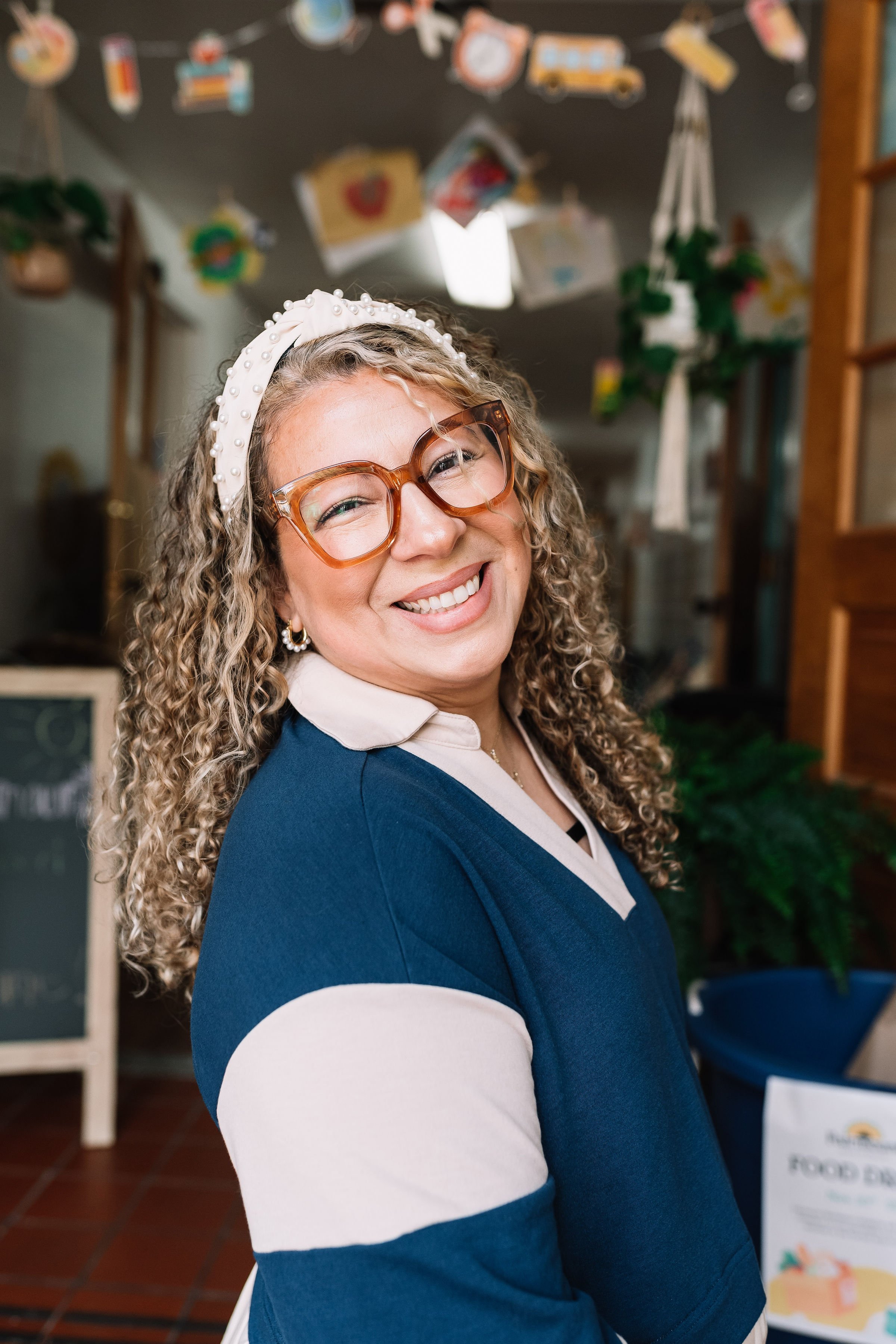 A woman with curly dark hair, glasses, and a beard, sitting indoors with a plant in the background.