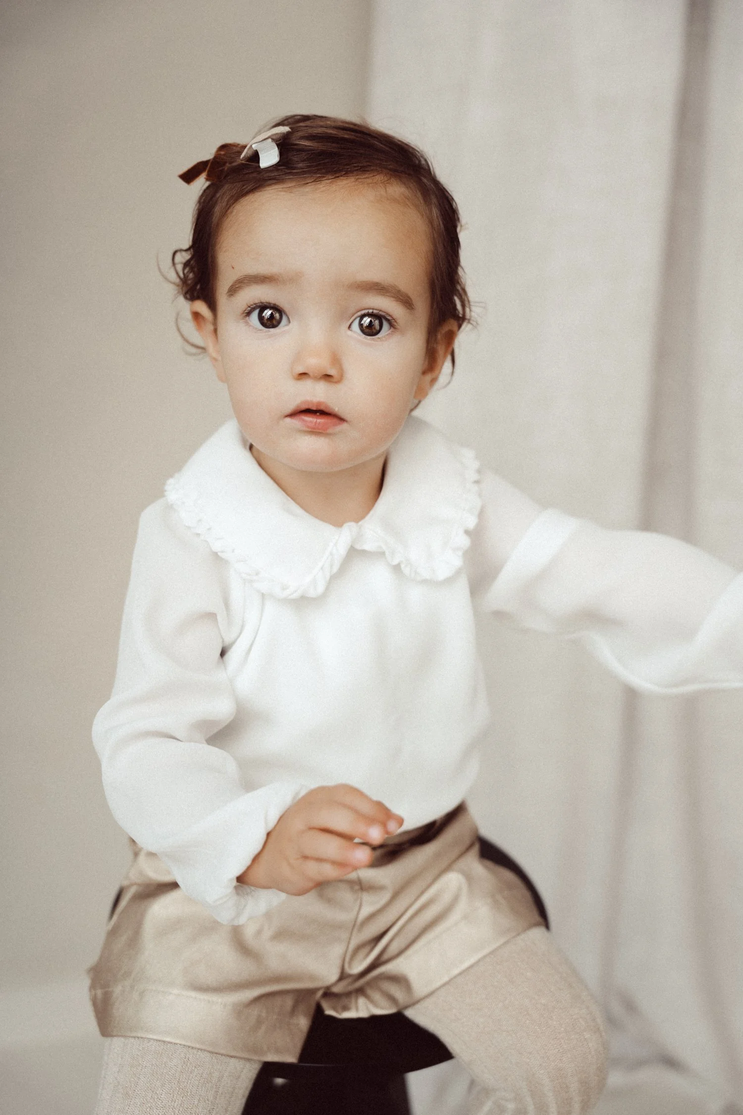A young girl with curly brown hair, wearing a white long-sleeve top with a ruffled collar and beige shorts, sitting on a black stool indoors with a neutral background.