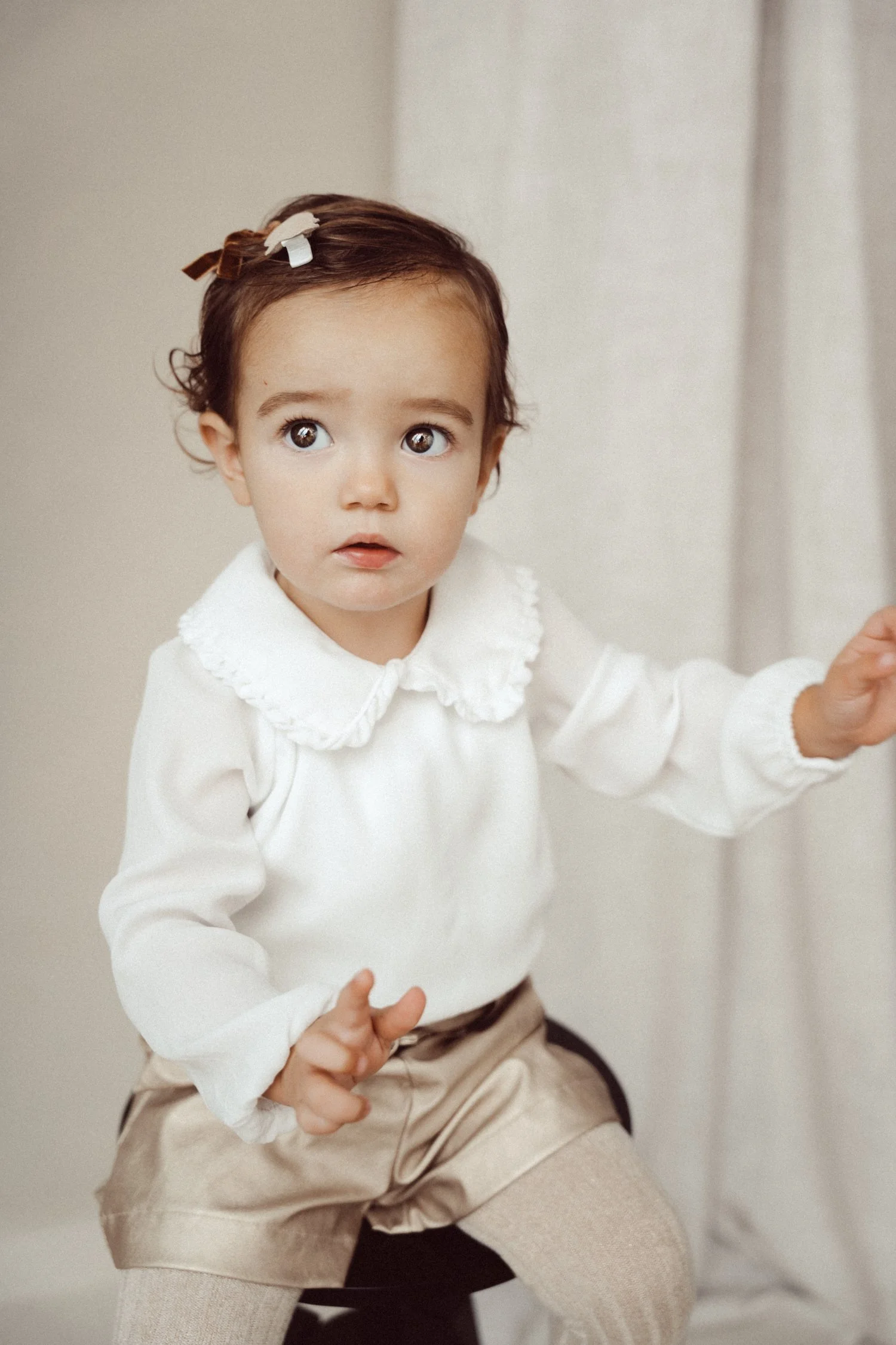 A young girl with brown hair, wearing a white shirt with a ruffled collar and beige shorts, sitting on a chair with a surprised or curious expression.