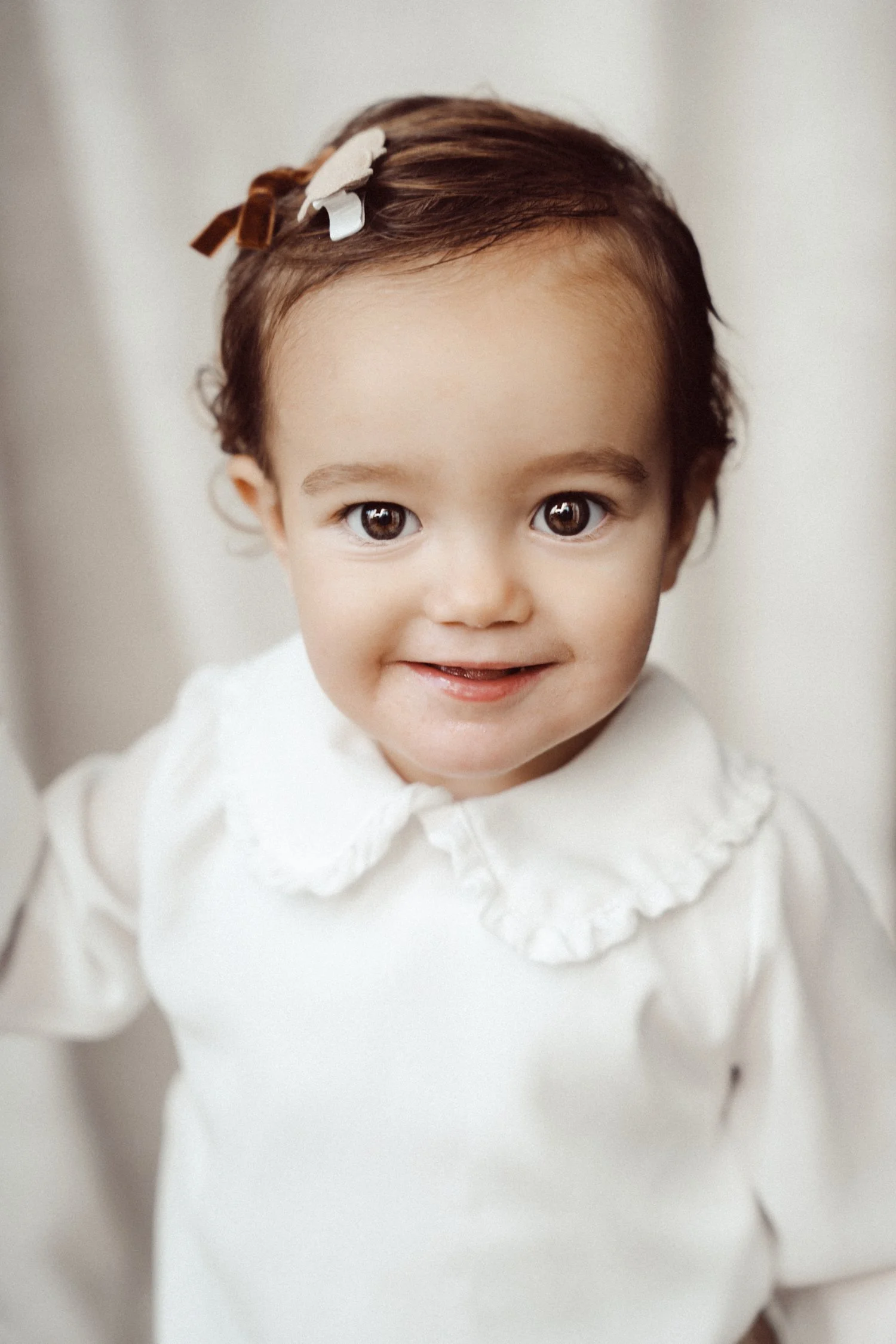 Close-up of a young girl with big brown eyes and dark brown hair, wearing a white dress with a ruffled collar, smiling at the camera.