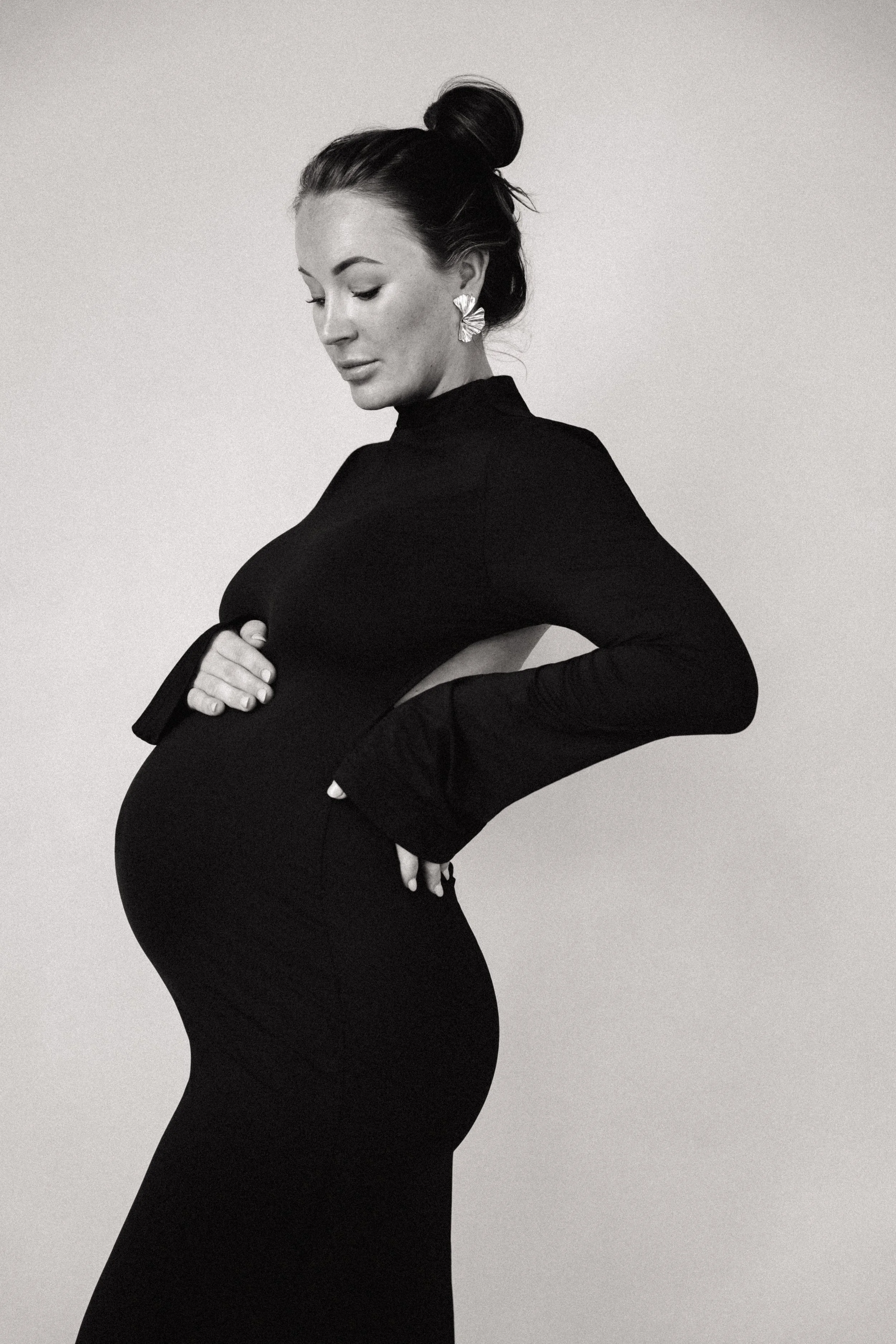 Black and white photo of a pregnant woman with her eyes closed, dressed in a long black dress with cut-out sides, posing against a plain background.