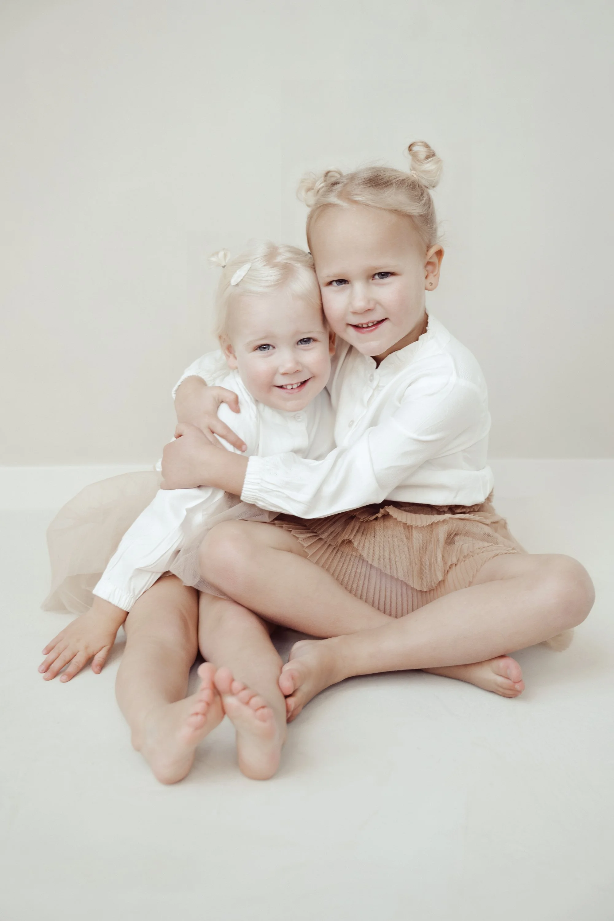 Two young girls sitting on the floor, hugging, and smiling at the camera, with one girl wearing a white shirt and the other a cream-colored skirt and blouse.