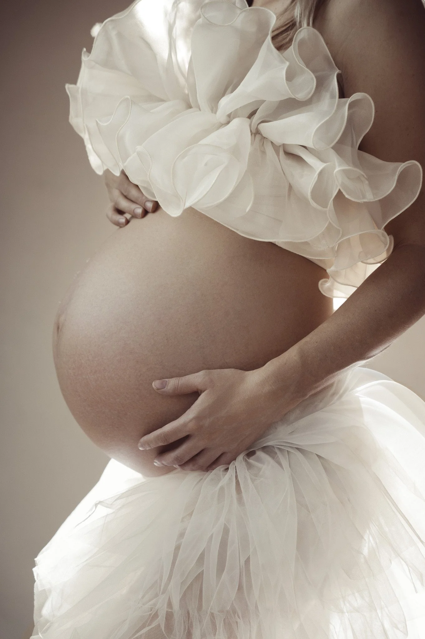 Black and white photo of a pregnant woman in a ruffled dress, looking to the side with her eyes closed, holding her belly with one hand and her shoulder with the other.