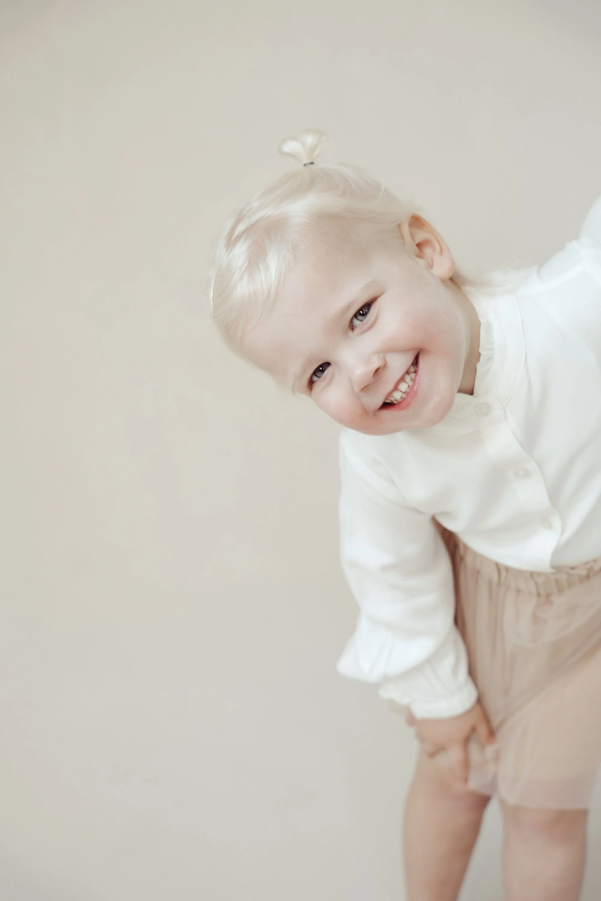 A smiling young girl with blonde hair, wearing a white top and beige shorts, leaning forward with her hand on her knee, against a plain light-colored background.