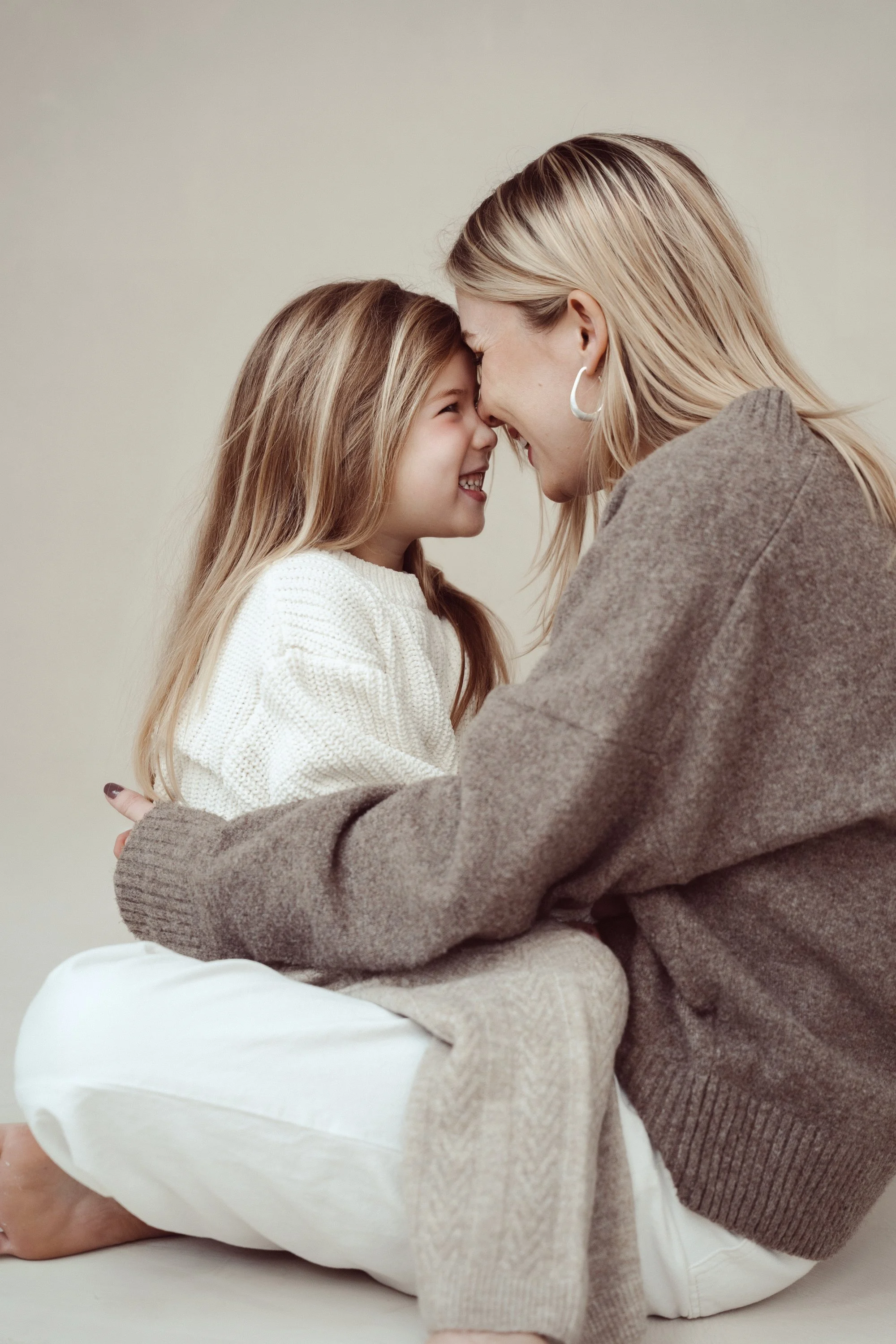 A woman and a young girl sitting crossed-legged on the floor, touching foreheads and smiling at each other affectionately.