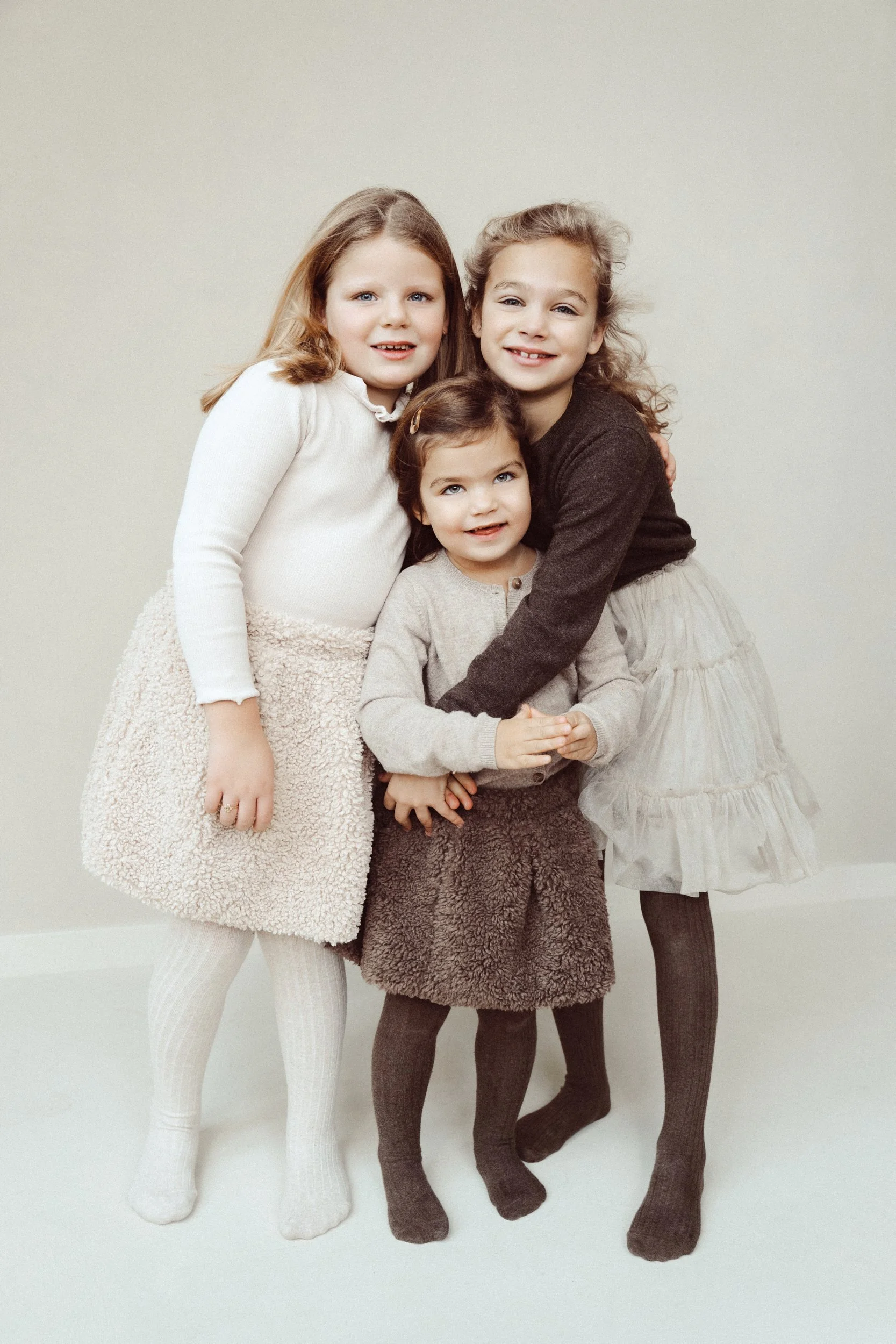 Four young girls with light skin and curly and straight hair, dressed in cozy winter clothes, standing together and smiling in front of a plain, light-colored background.