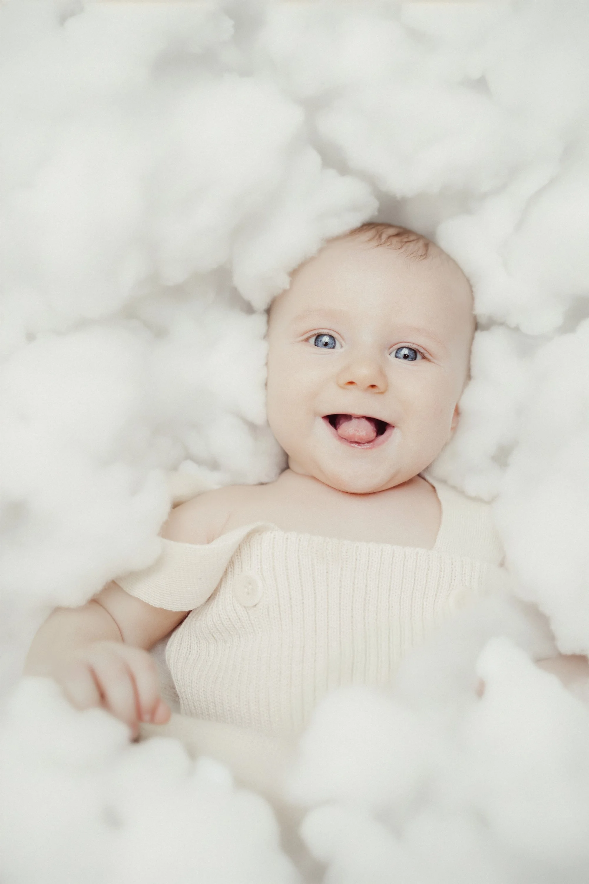 A smiling baby lying on fluffy white clouds, wearing a cream-colored sleeveless top.