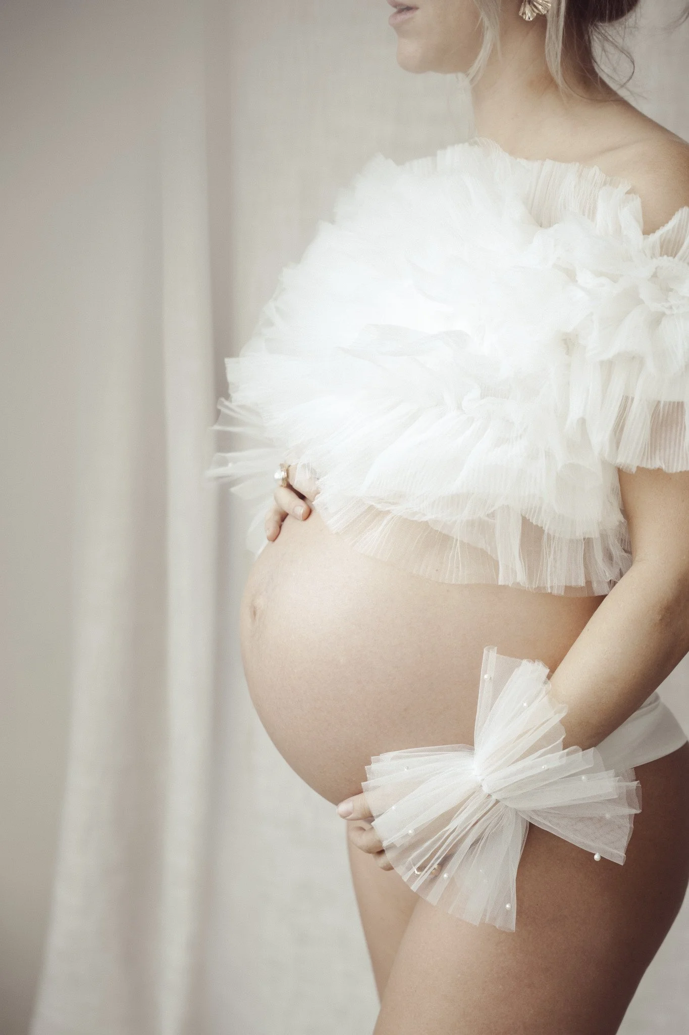 Black and white photo of a pregnant woman in a ruffled dress, looking to the side with her eyes closed, holding her belly with one hand and her shoulder with the other.