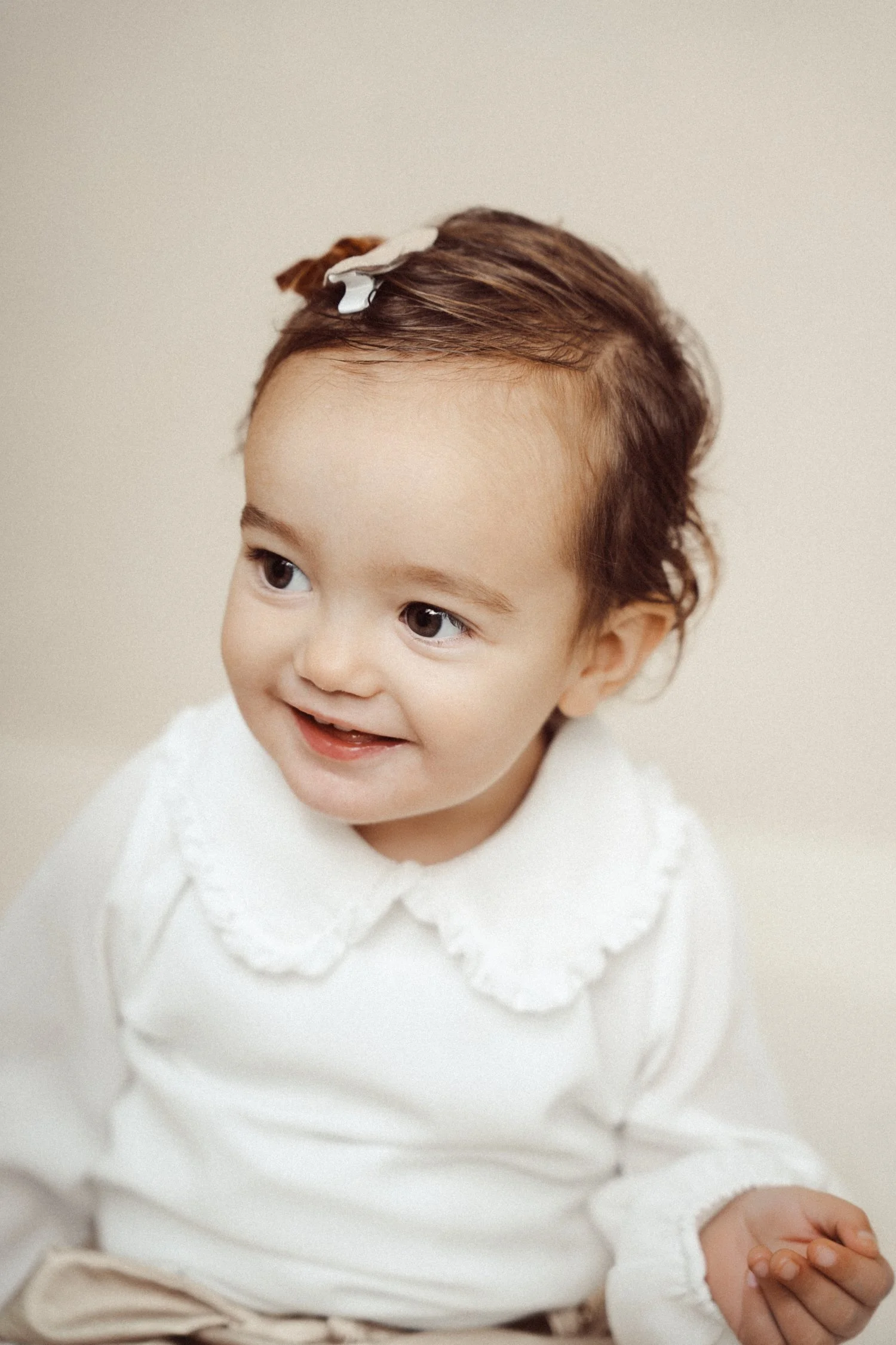 A smiling young girl with brown hair, wearing a white blouse with a lace collar, sitting indoors against a neutral background.