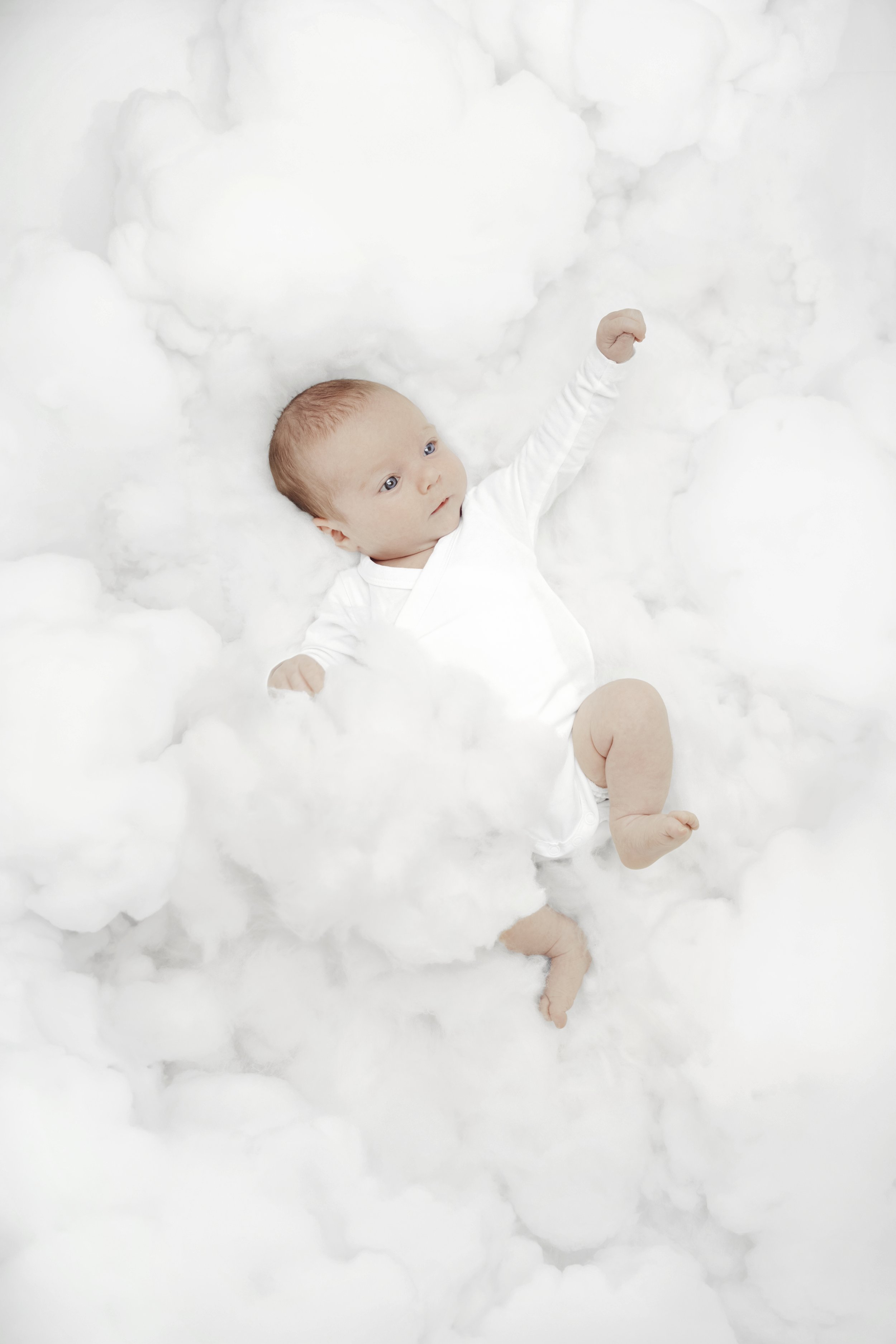 A baby lying among fluffy white clouds, reaching upward with one arm.