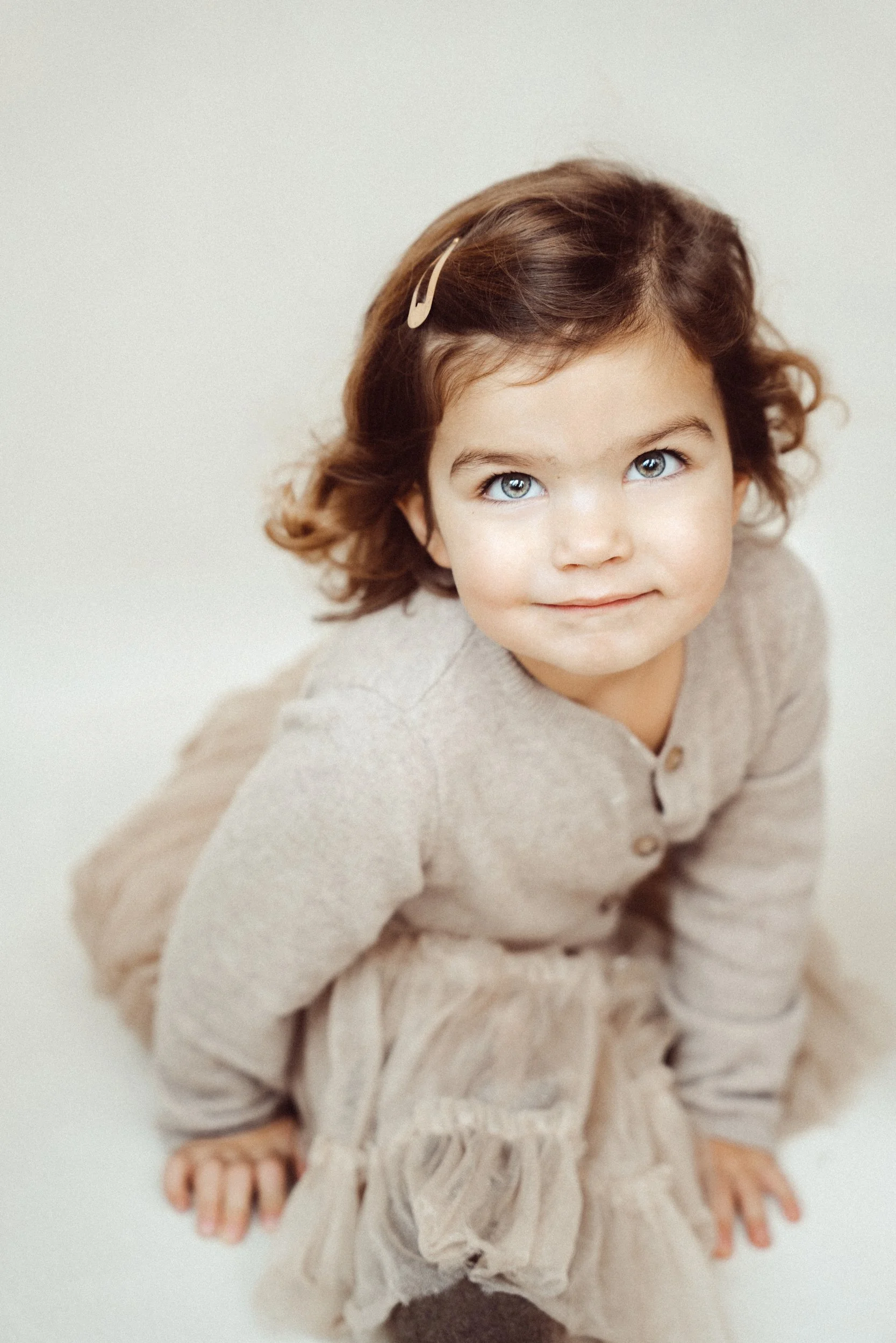A young girl with brown, curly hair, blue eyes, and wearing a beige sweater and matching skirt, is looking at the camera with a gentle smile.