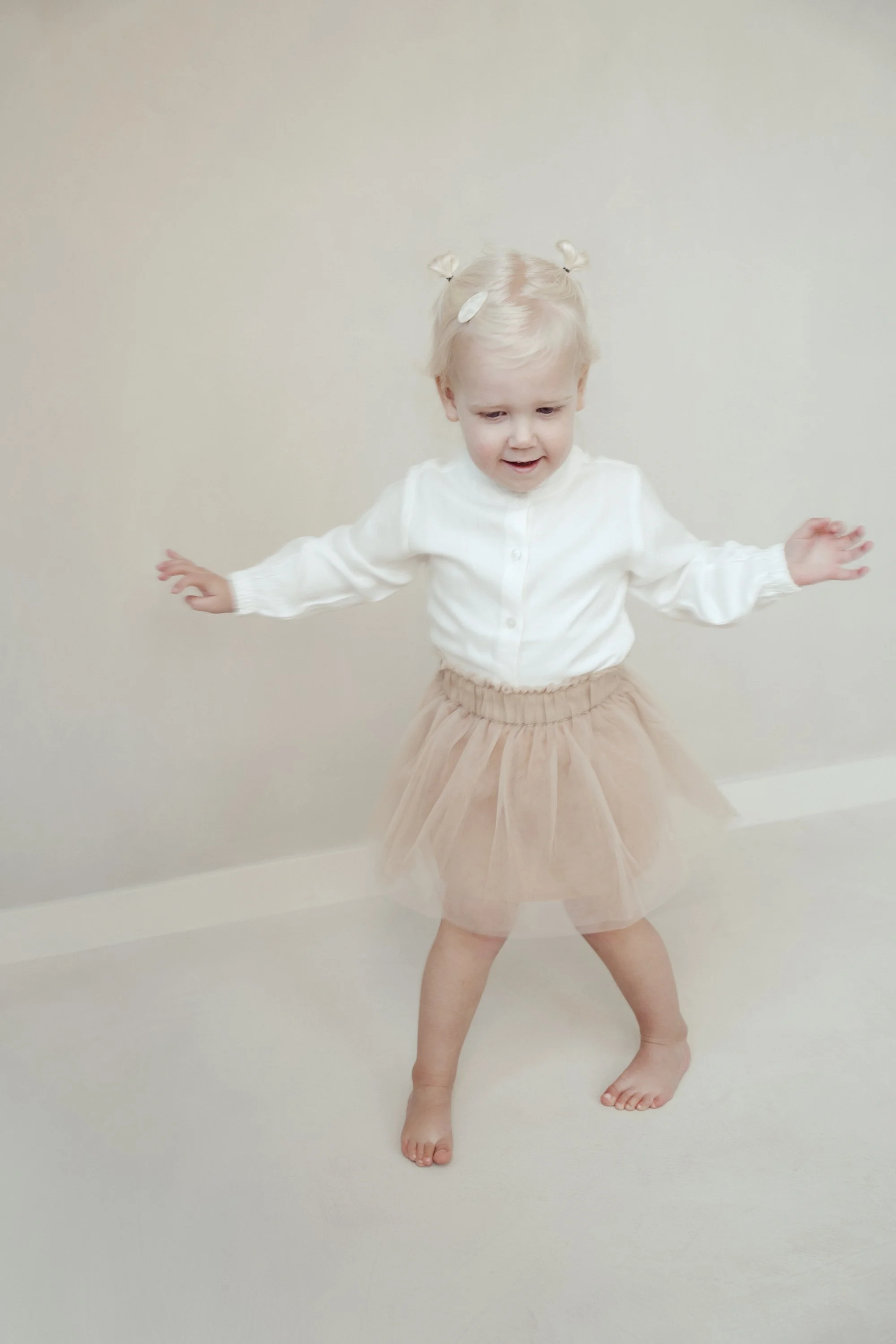 A young girl with blonde hair, wearing a white long sleeve top and a beige tulle skirt, standing on a cream-colored carpet. She has her arms spread out and appears to be balancing or dancing against a plain beige wall.