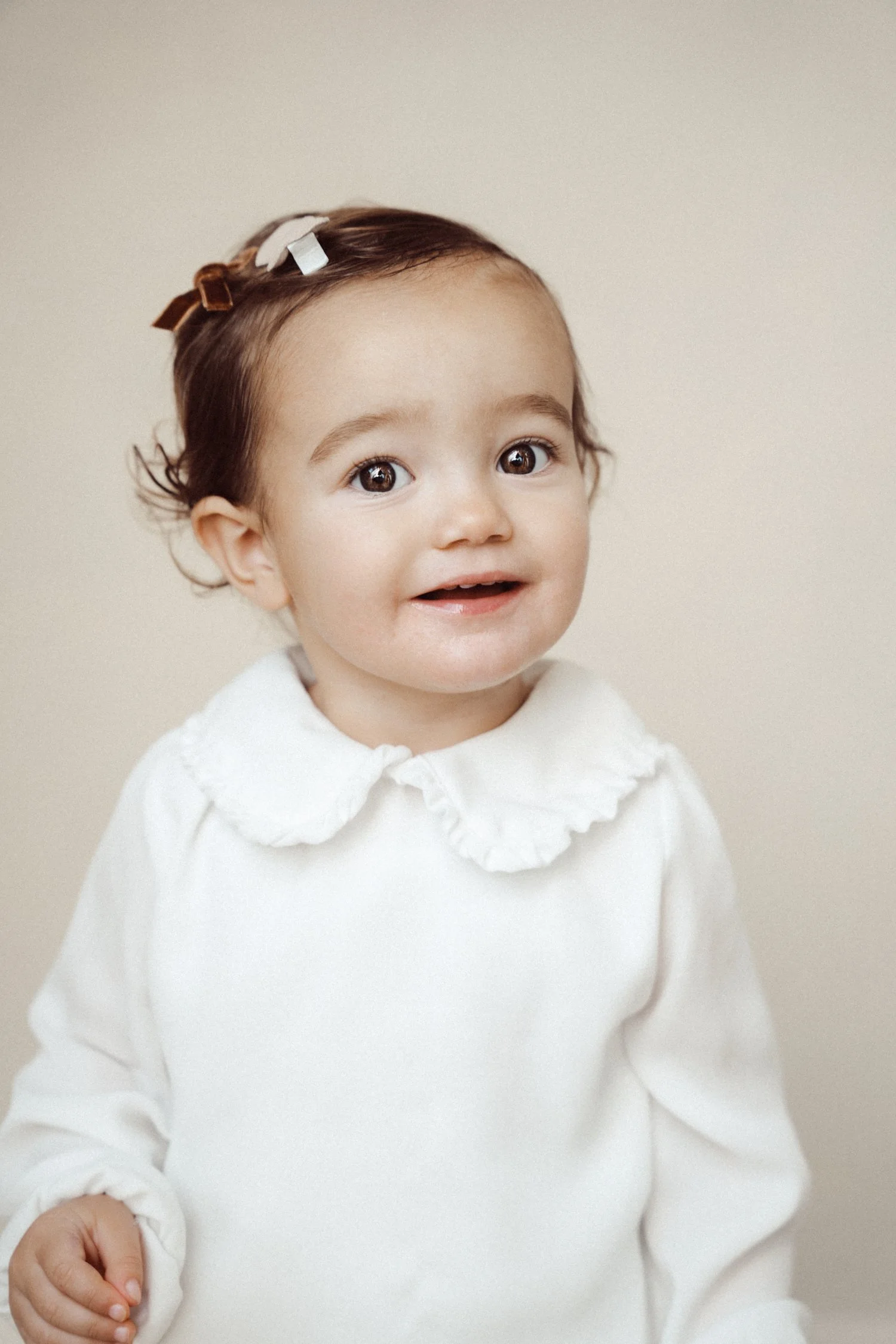 A young girl with curly brown hair, large brown eyes, and a happy expression, wearing a white top with a ruffled collar and a headband with white and brown bows, standing against a beige background.