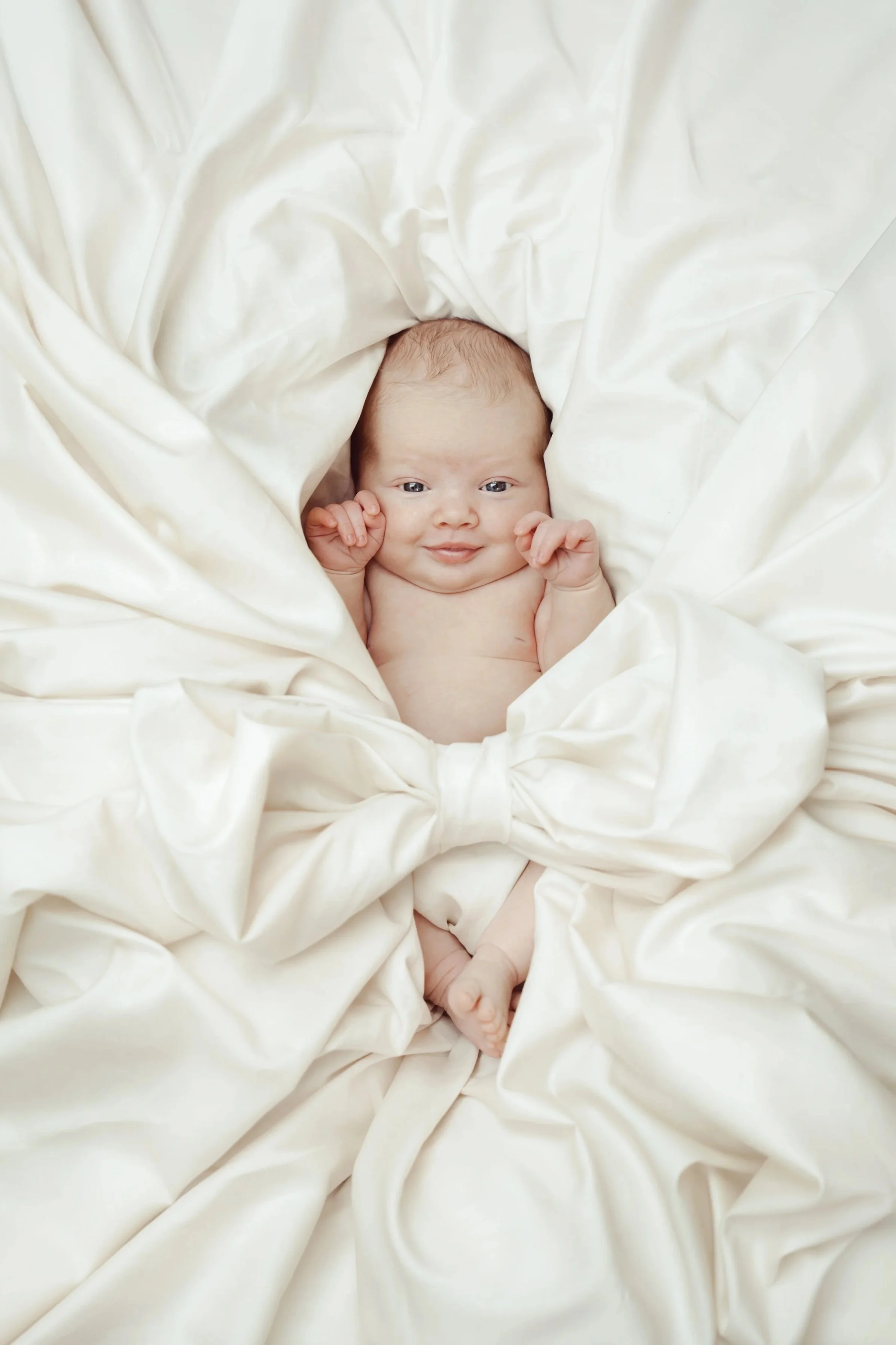 A smiling baby lying on a bed among white, crumpled sheets.