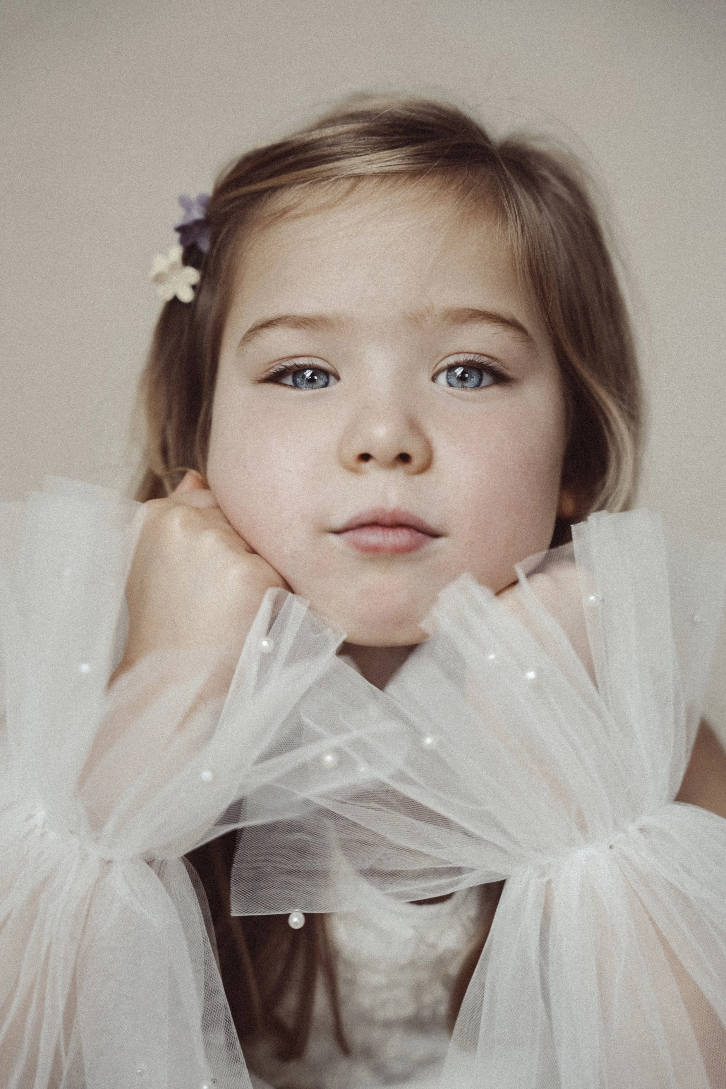 A young girl with blue eyes and light brown hair, wearing a white tulle dress with pearl embellishments, resting her chin on her hands, and looking directly at the camera.