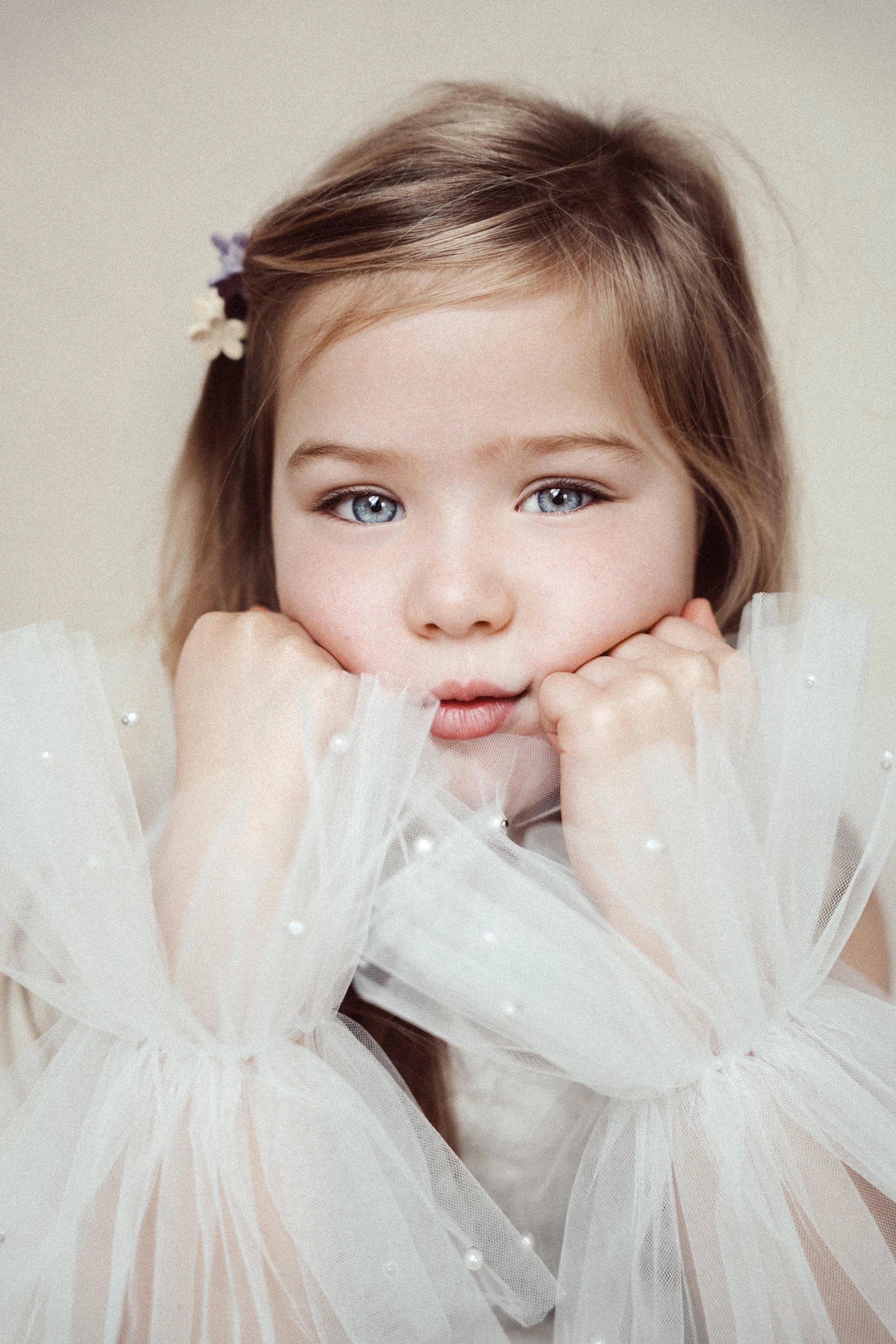 A young girl with blue eyes and light brown hair, wearing a white tulle dress with pearl embellishments, resting her chin on her hands, and looking directly at the camera.
