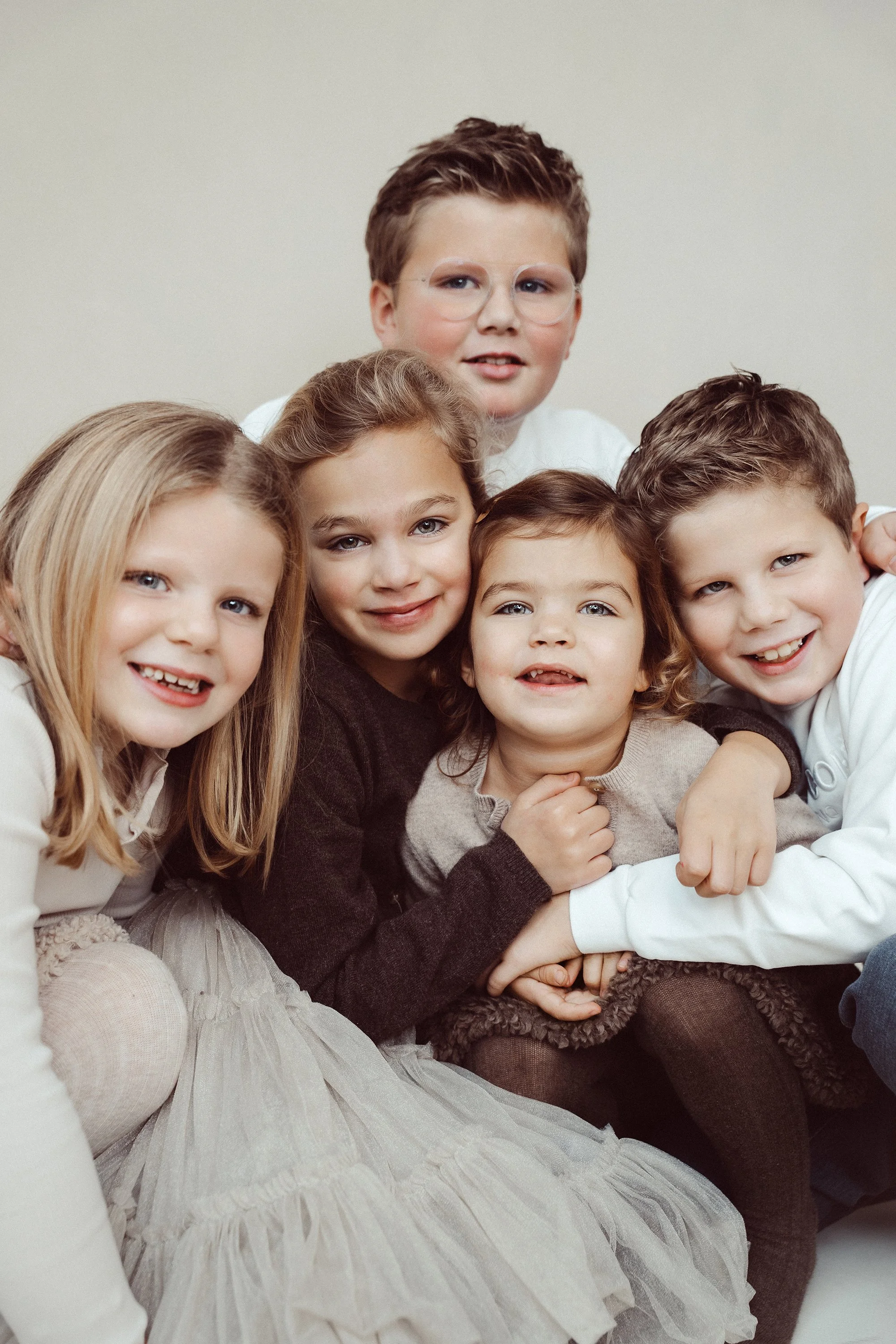 Group of five children sitting closely together, smiling at the camera, against a plain neutral background.