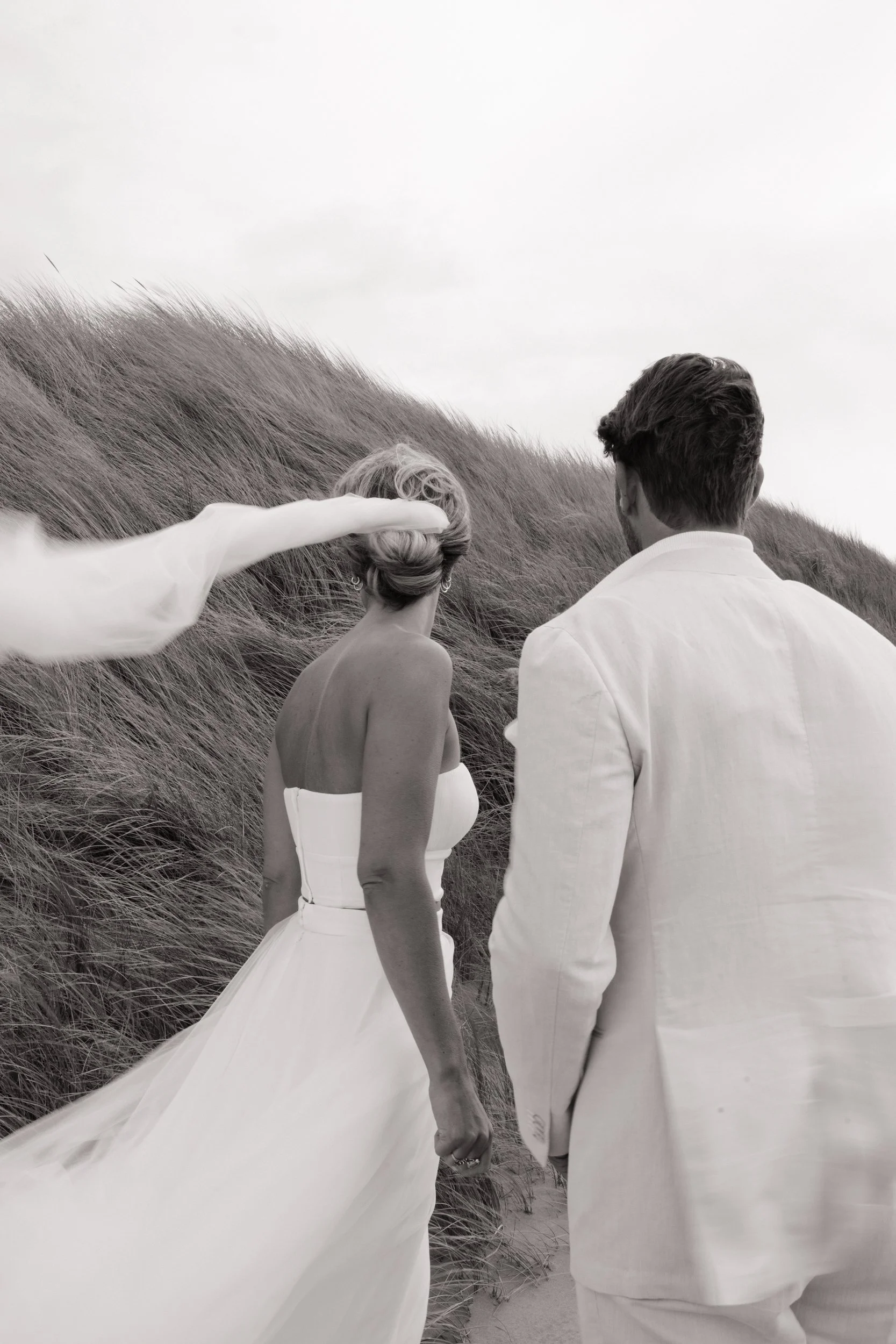 A black and white photo of a man and woman in wedding attire at the beach, with a person saluting them from the side, and tall grass on dunes in the background.