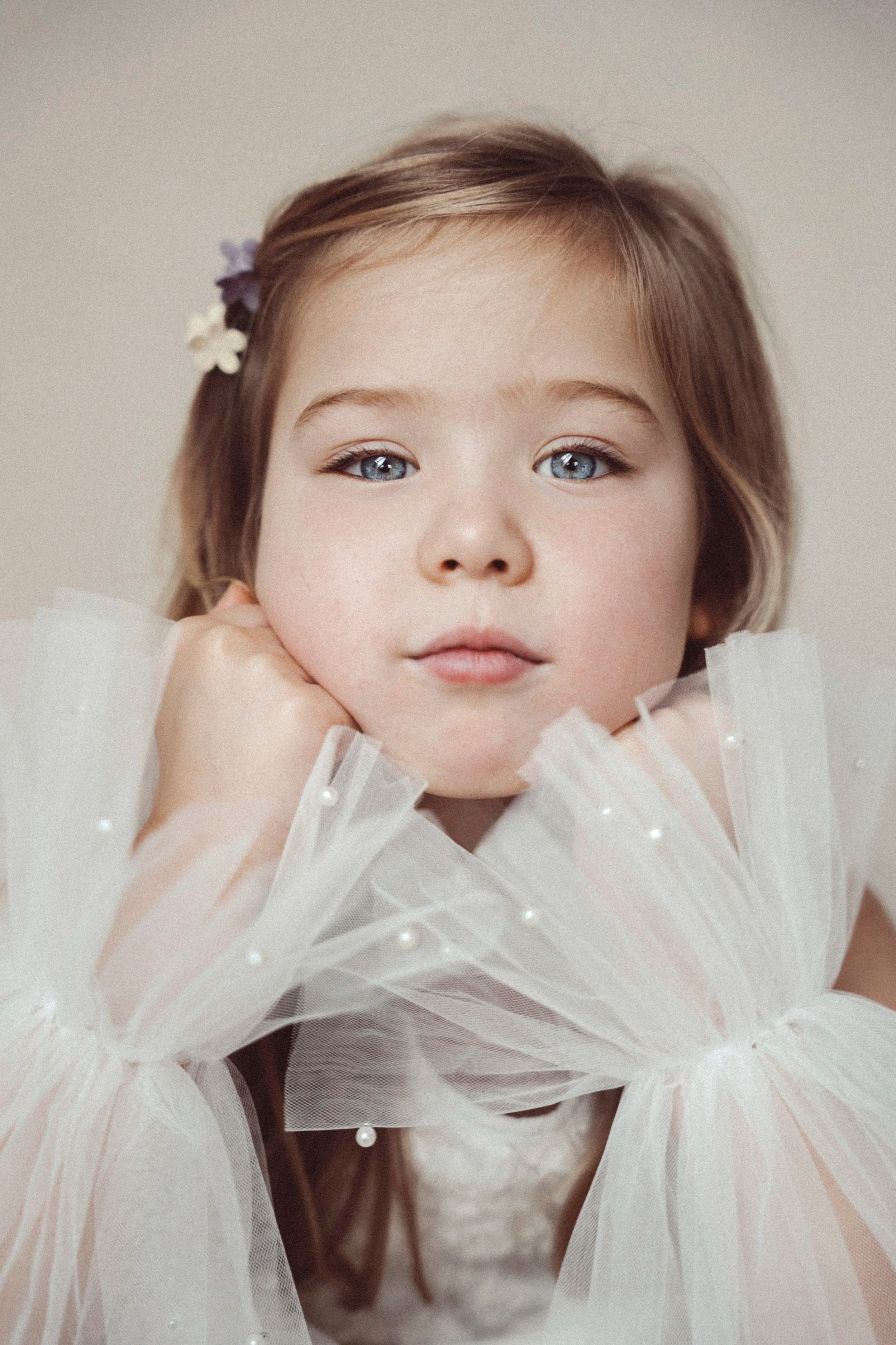 A young girl with blue eyes and light brown hair, wearing a white tulle dress with pearl embellishments, resting her chin on her hands, and looking directly at the camera.