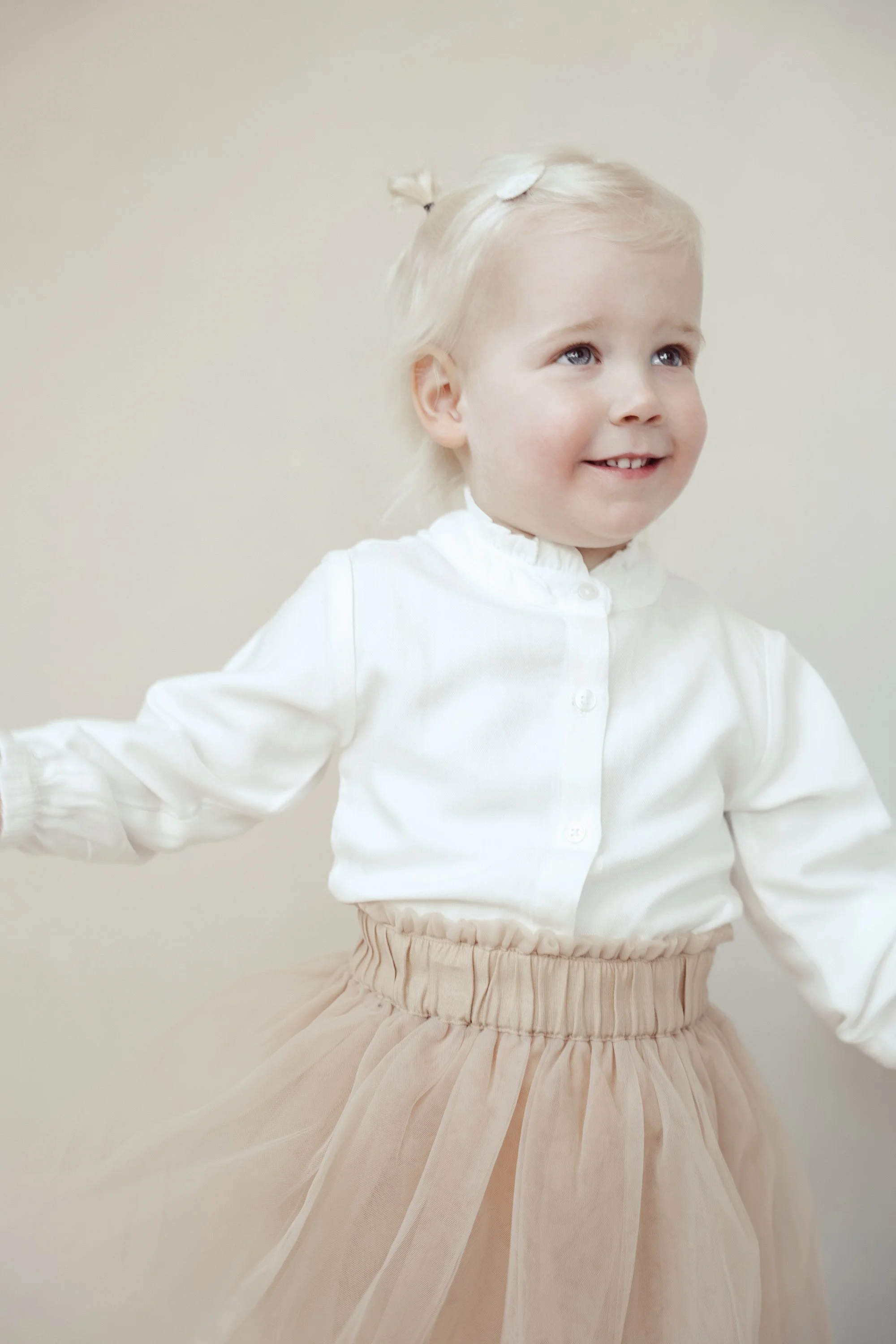 A young girl with blonde hair tied with a small bow, wearing a white blouse and a beige tulle skirt, smiling and looking to the side against a plain light-colored background.