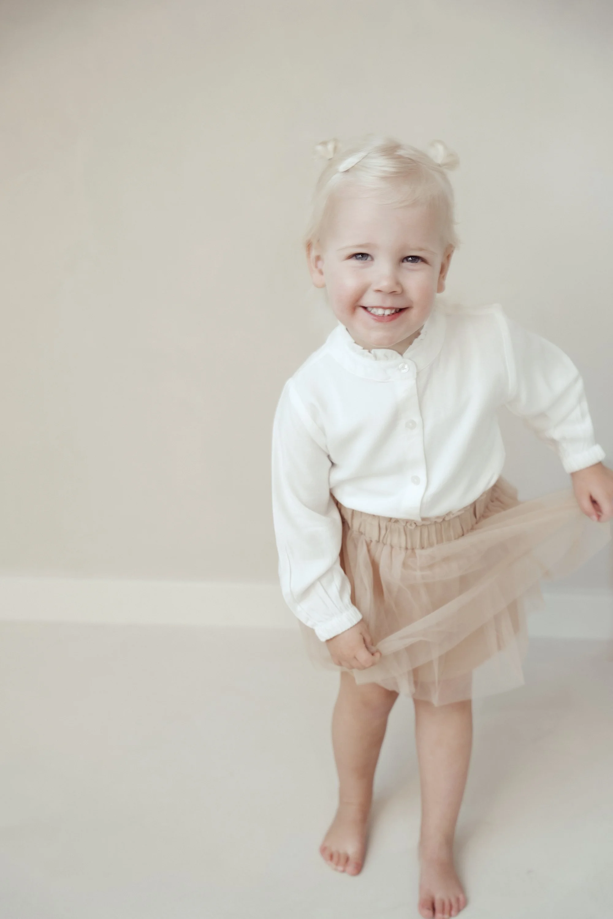 A young girl with blonde hair styled in two small buns, wearing a white long-sleeve blouse and a beige tulle skirt, smiling and standing barefoot against a plain light-colored wall.