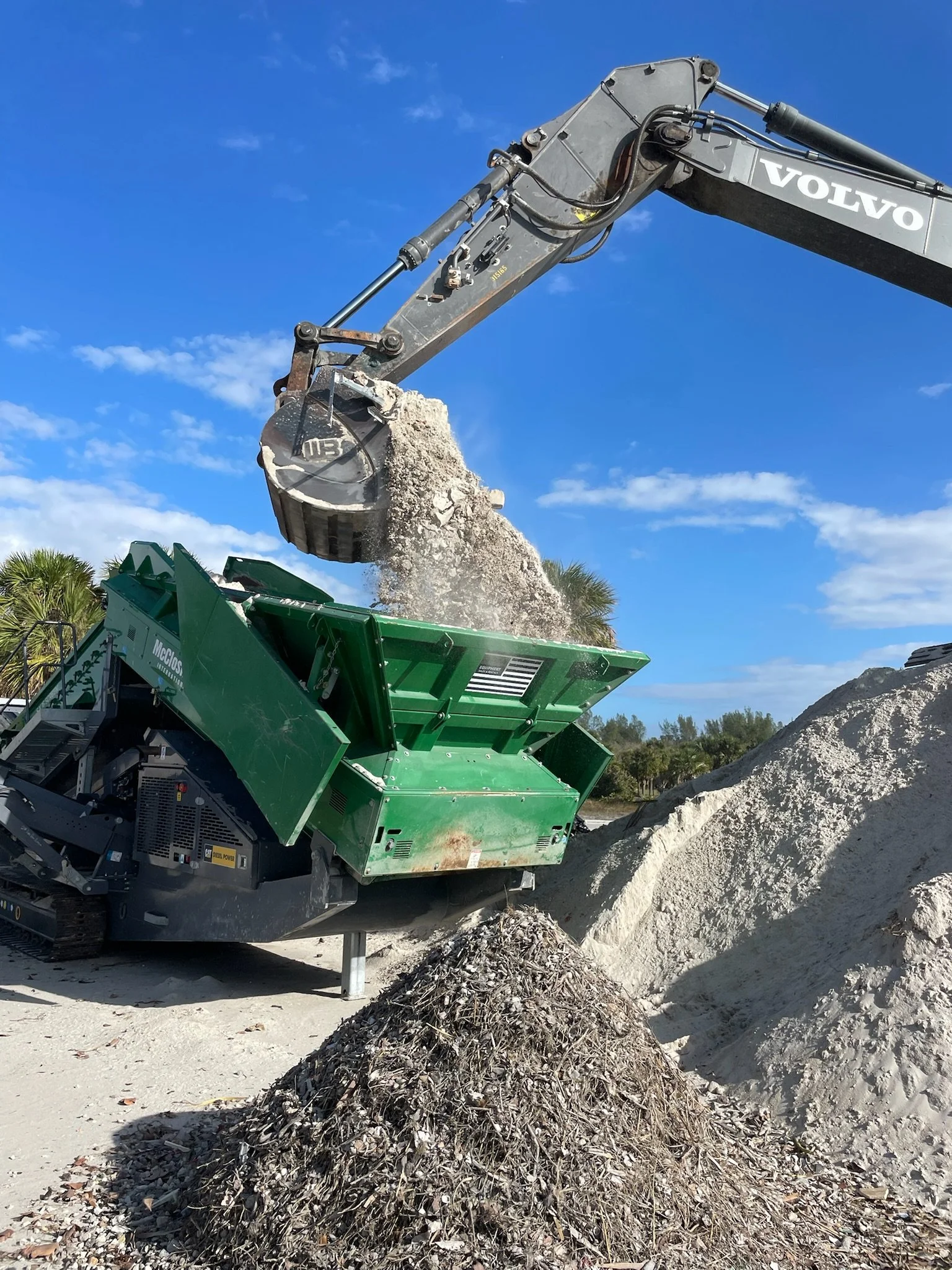 A large green and gray excavator with the brand 'Volvo' is dumping a load of tree branches and debris onto a pile of similar material at an outdoor work site with a clear blue sky and some trees in the background.