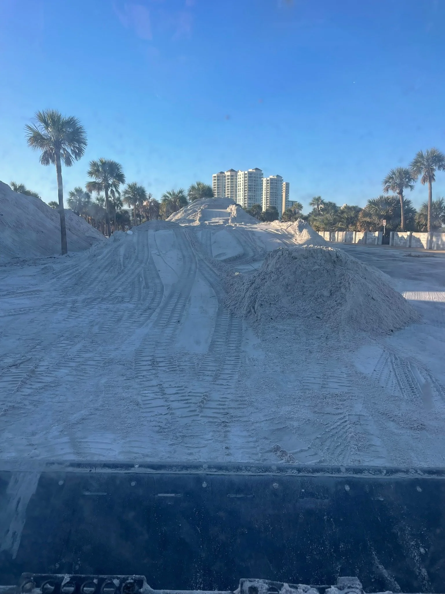 Photo taken from inside a vehicle showing a snowy road with tire tracks, snow piles on the sides, and palm trees with modern high-rise buildings in the background under a blue sky.