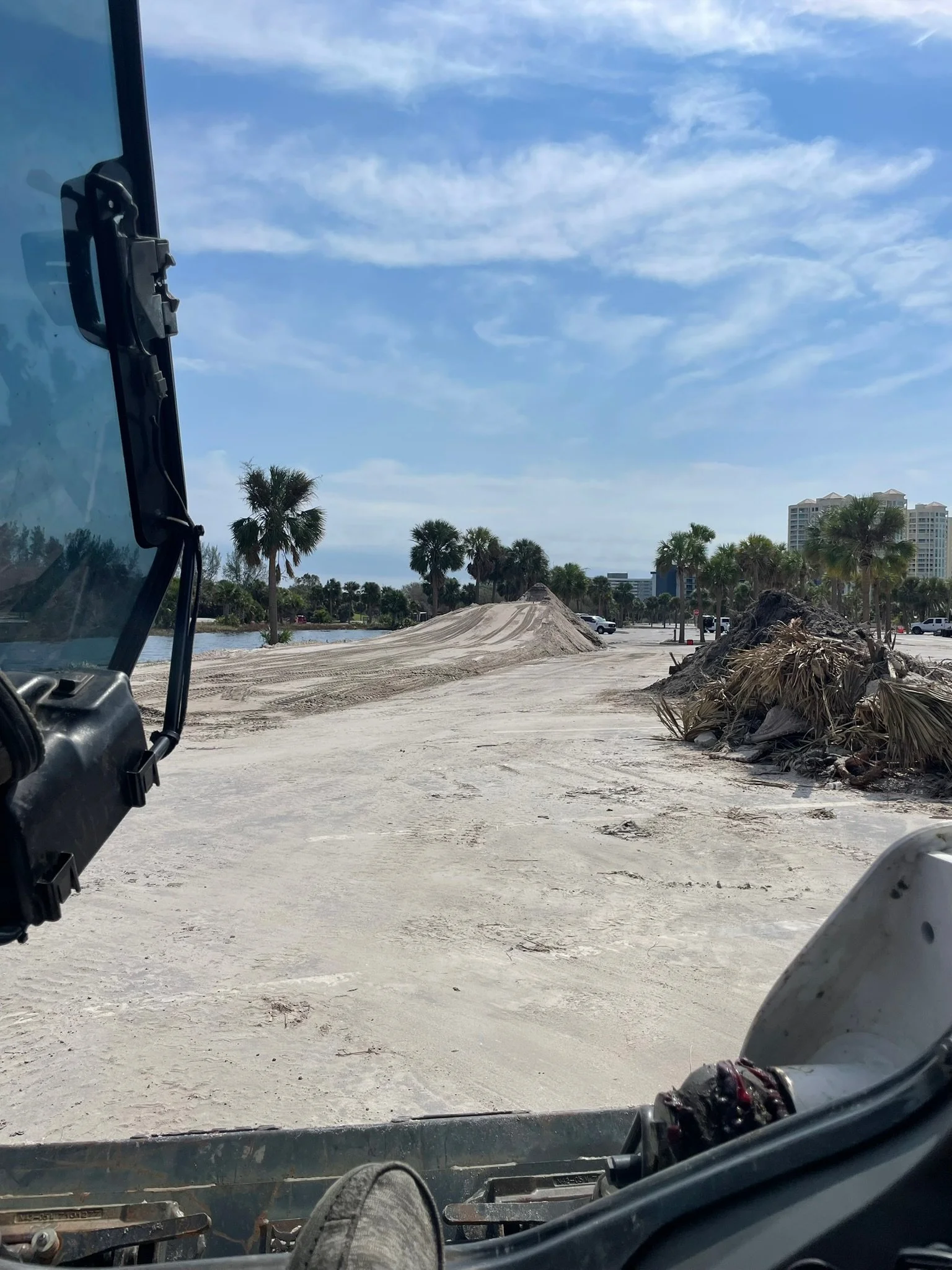 View from inside a construction vehicle looking over a sandy area with piles of debris, palm trees, and buildings in the distance under a partly cloudy sky.