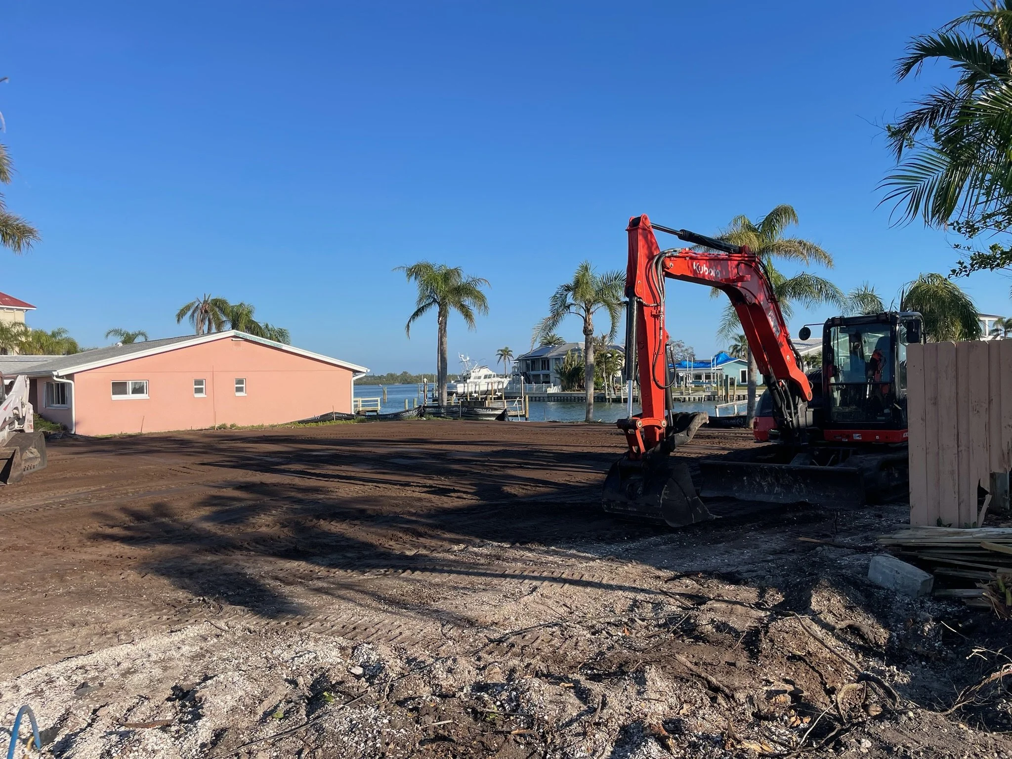 A construction site near a waterway with a pink house, palm trees, and a boat in the background. A red excavator is on the site, which has a dirt ground with tire tracks.