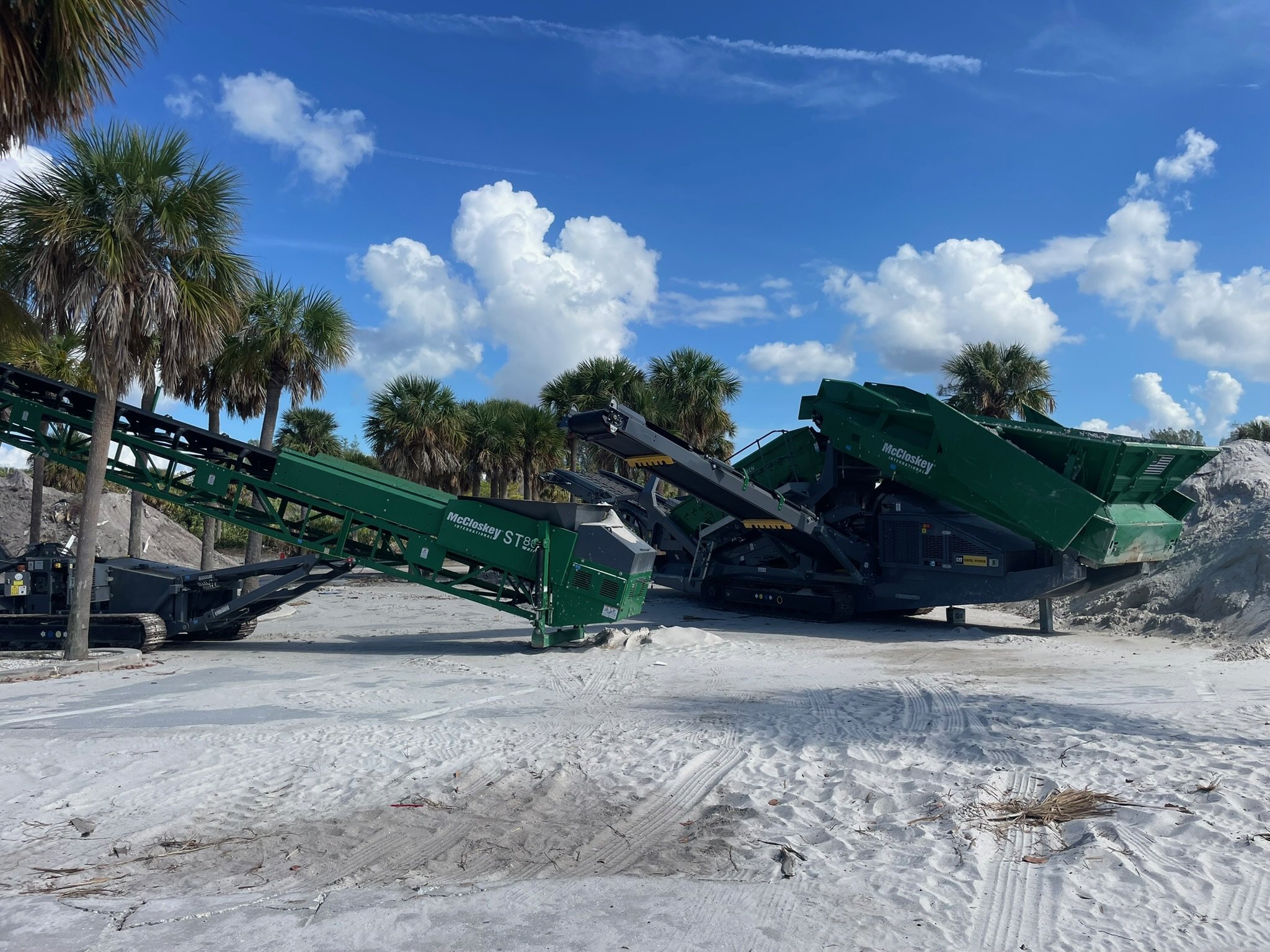 Construction equipment on a sandy beach with palm trees and a blue sky with clouds.