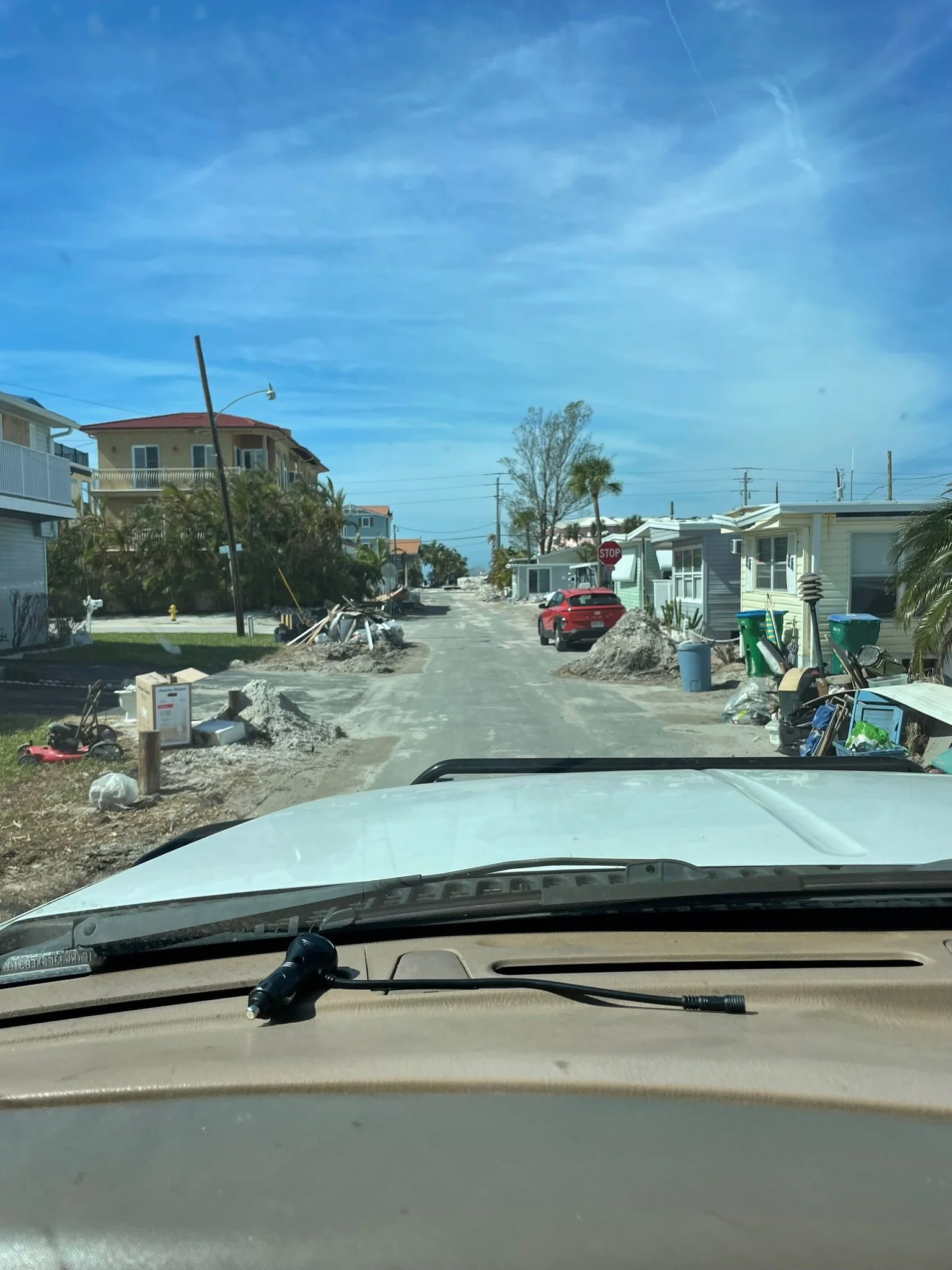 Residential street with houses, debris, construction materials, and a red car, under a blue sky with clouds.