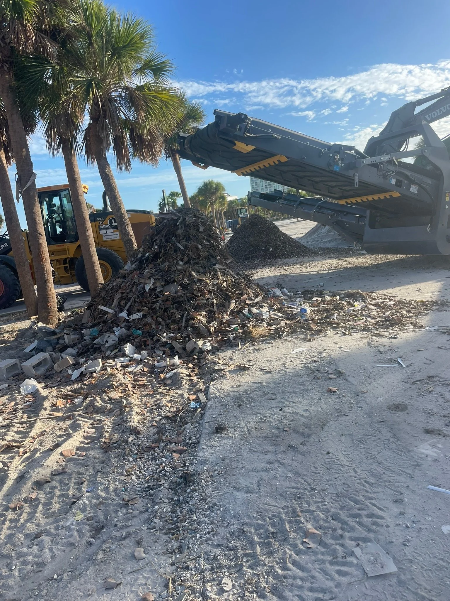 A construction site with a large excavator lifting debris, piles of rubble and trash, and several palm trees under a blue sky with scattered clouds.