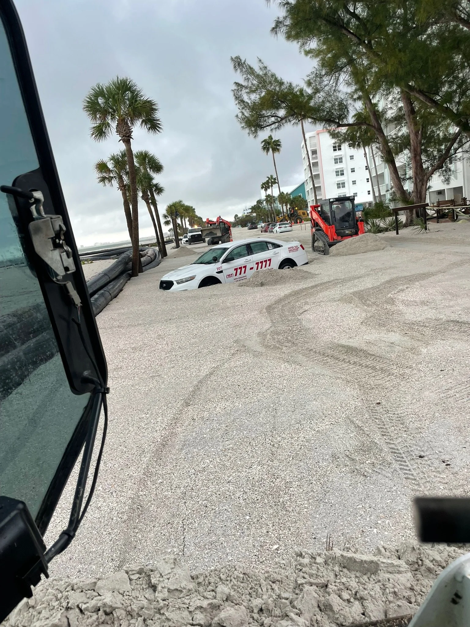 A construction site near a beach with palm trees and multi-story buildings in the background. Construction equipment and vehicles are visible, including a small red excavator and a white car with red and black markings. Sand and gravel are on the gro