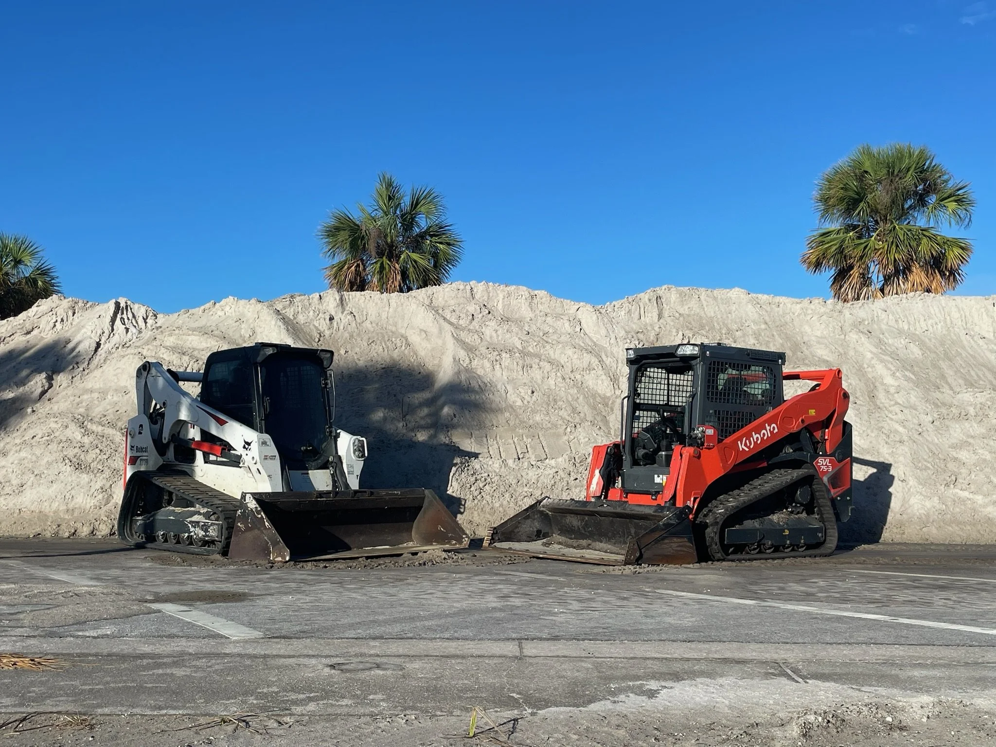 Construction equipment with a white compact loader and an orange compact tractor parked on a paved area in front of a dirt hill with palm trees and a clear blue sky.