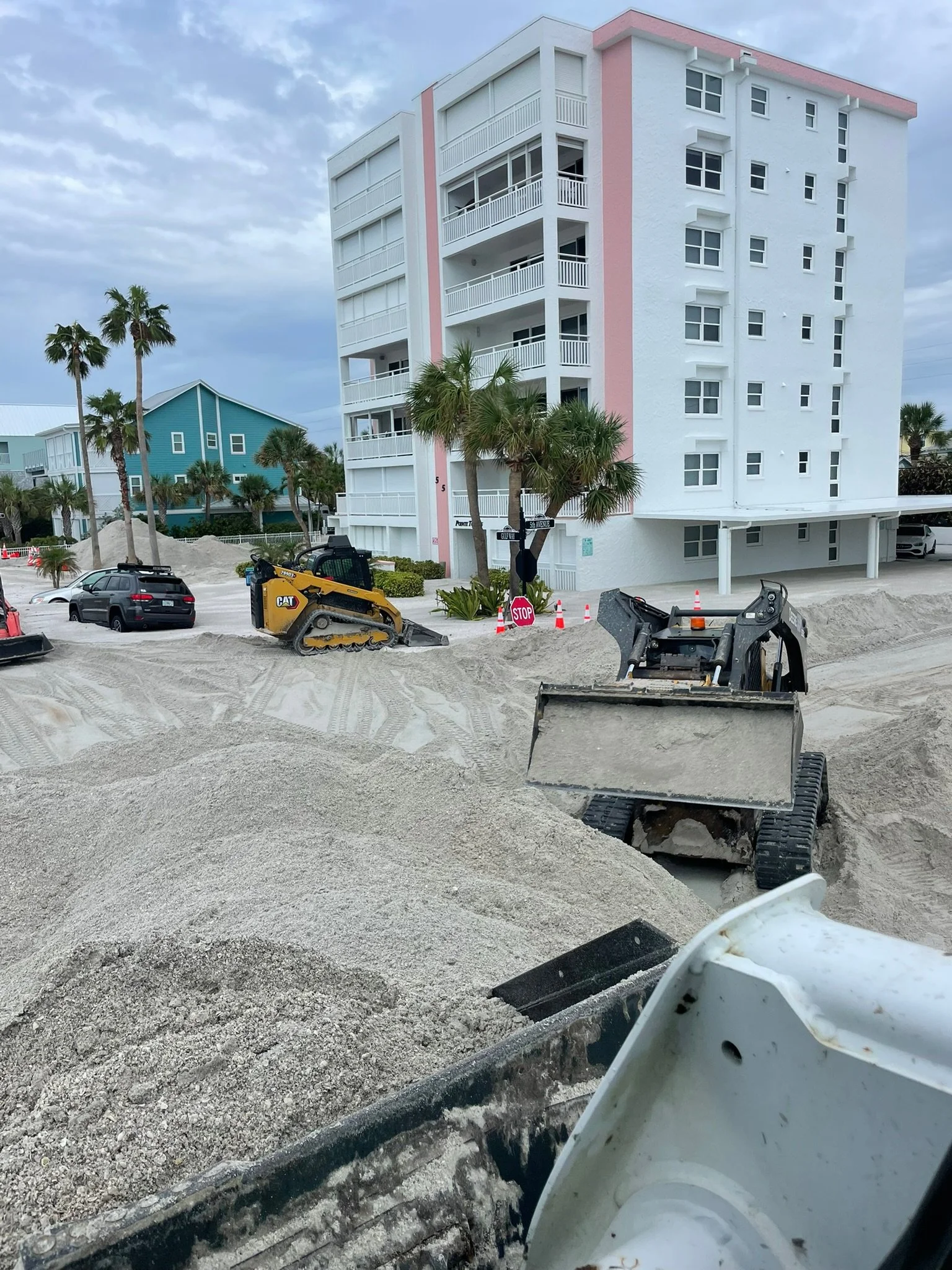 Construction site with sand, construction vehicles, and a tall white building with pink accents and balconies. Palm trees are in the background under cloudy sky.