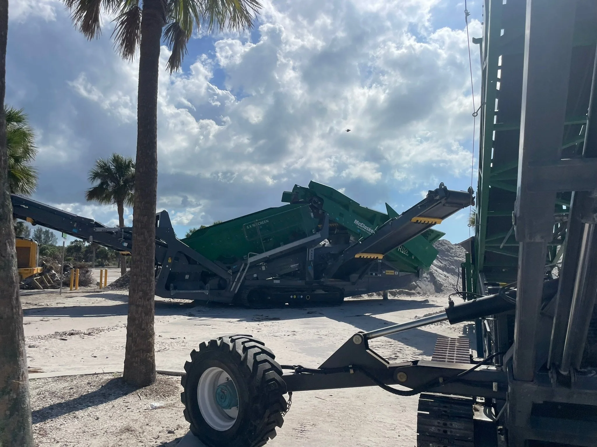 Construction site with a large green machine and palm trees under a cloudy sky.
