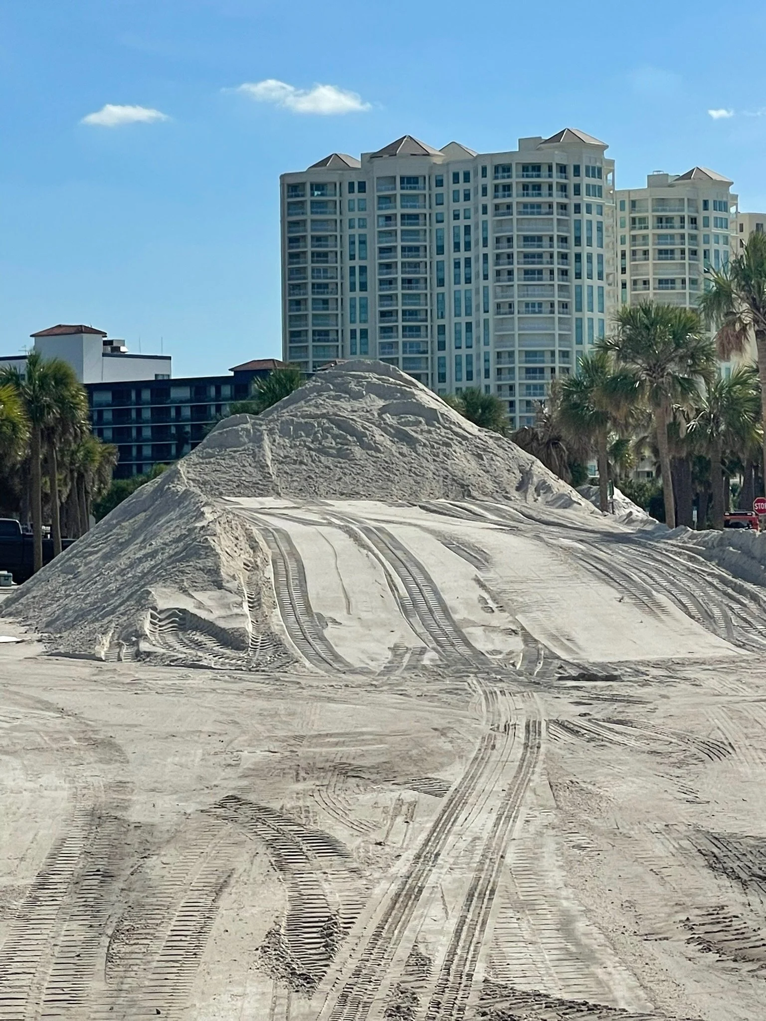 A sandy area with tire tracks and a mound of sand, with high-rise buildings and palm trees in the background under a blue sky with a few clouds.