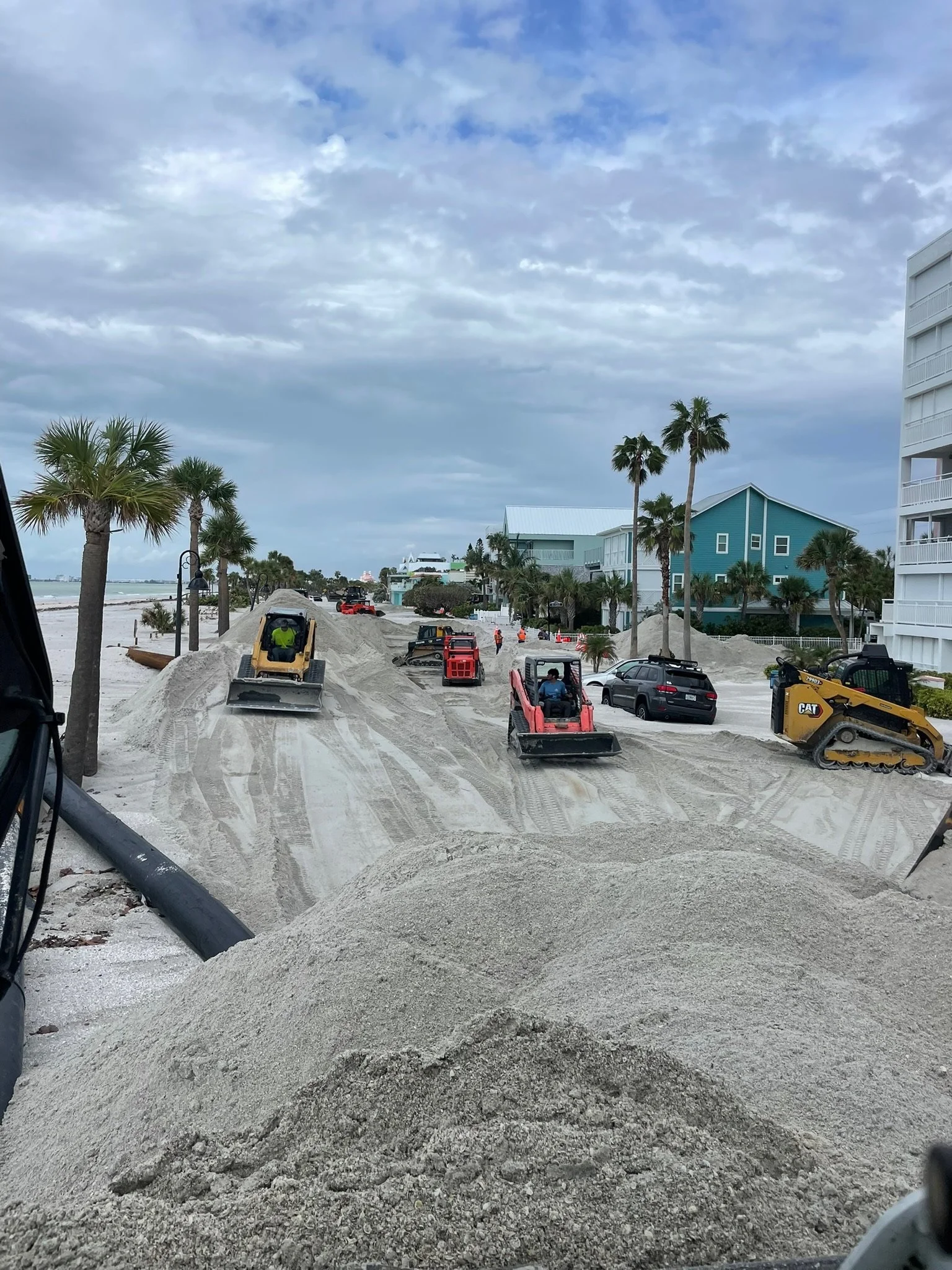 Beachside construction site with sand piles, construction machinery, construction workers, and nearby buildings with palm trees and overcast sky.