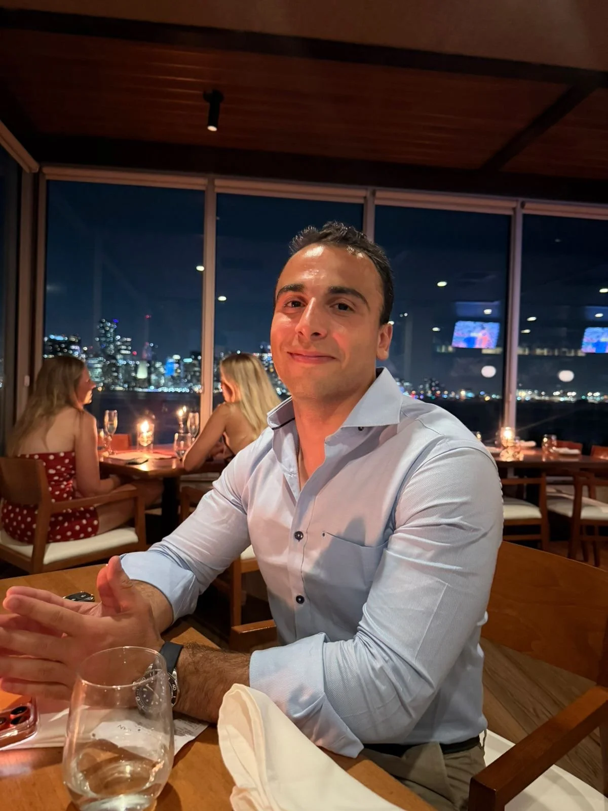 A man in a light blue dress shirt sitting at a restaurant table with a city skyline at night in the background, with two women sitting at another table.