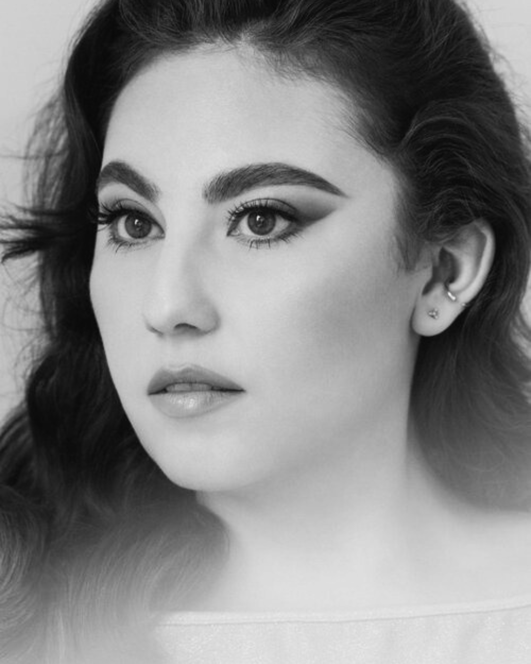 Black and white portrait of a woman with styled hair, winged eyeliner, and earrings, with the words 'MARIA CURIEL' overlaid.