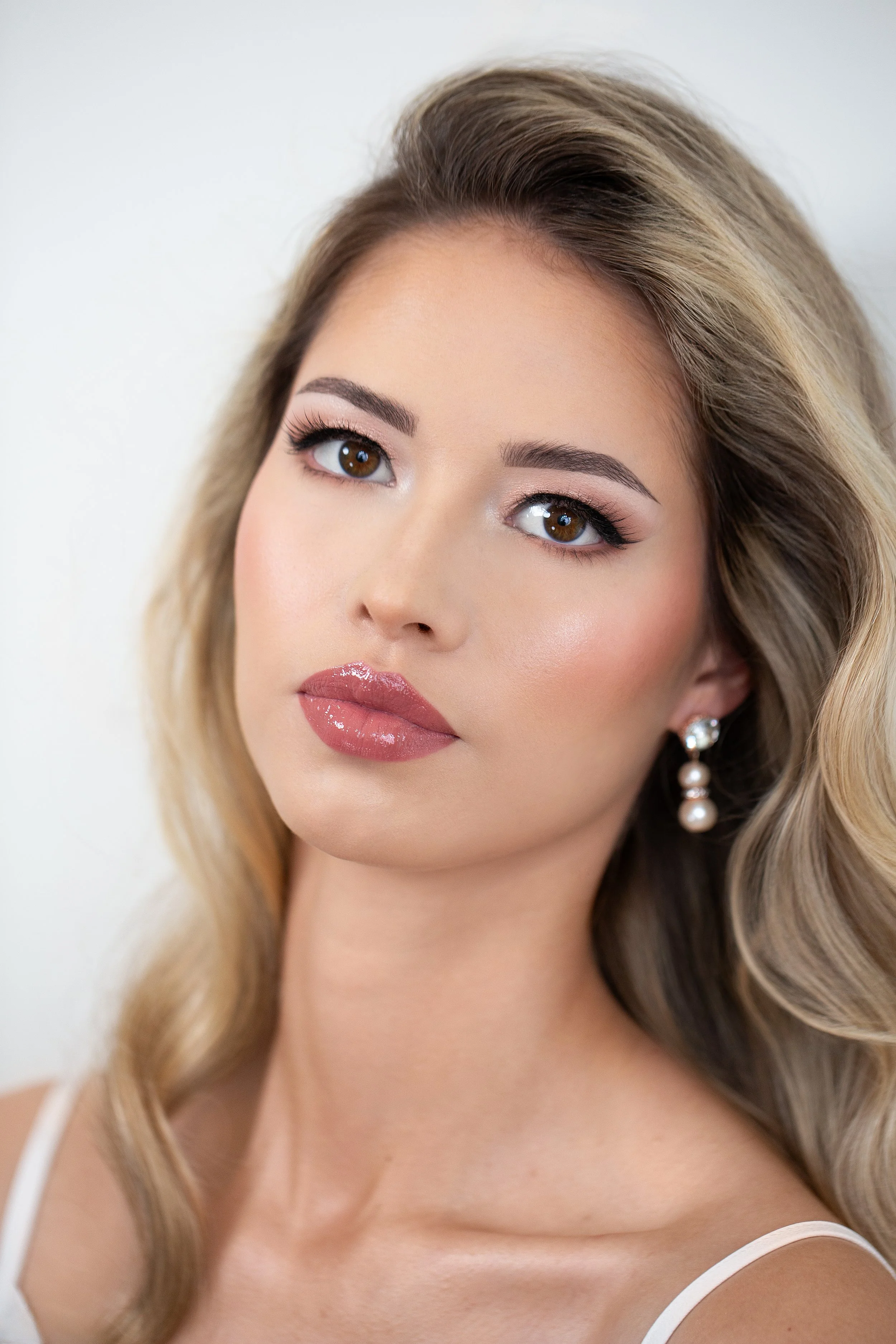 Close-up portrait of a woman with makeup and wavy blonde hair, wearing pearl earrings and a white top.
