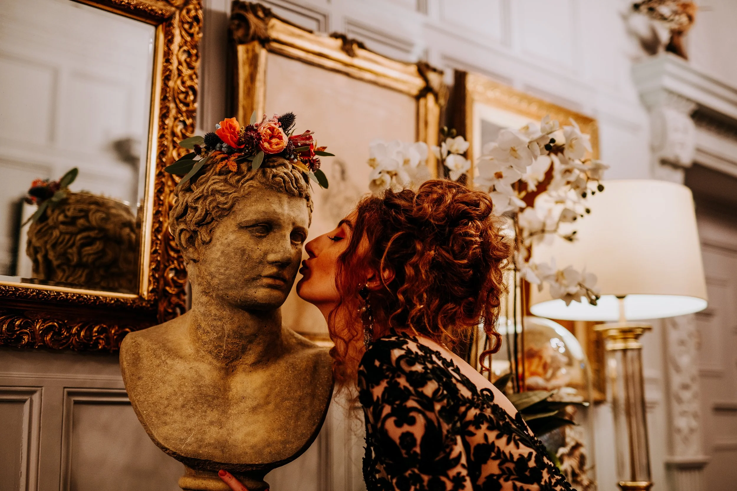A woman with curly red hair and earrings kisses a stone bust with a floral crown in an elegant, decorated room.