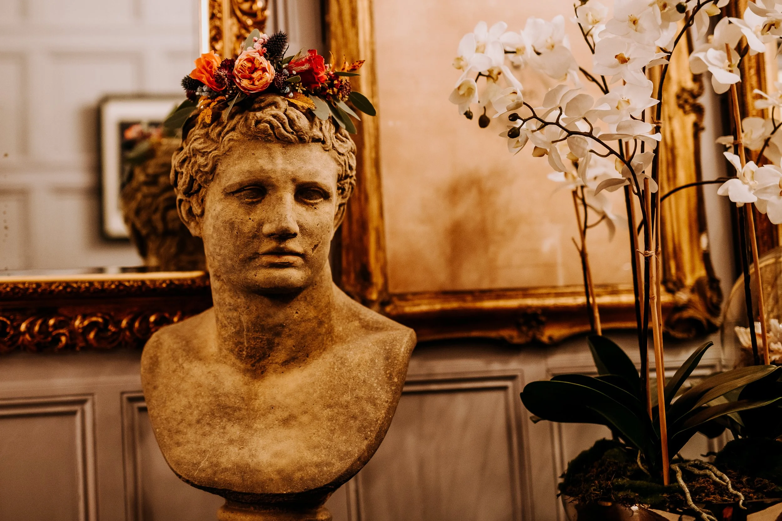 Ancient bust sculpture of a woman wearing a floral crown, placed in a luxurious antique setting with ornate gold-framed mirror and white orchids.