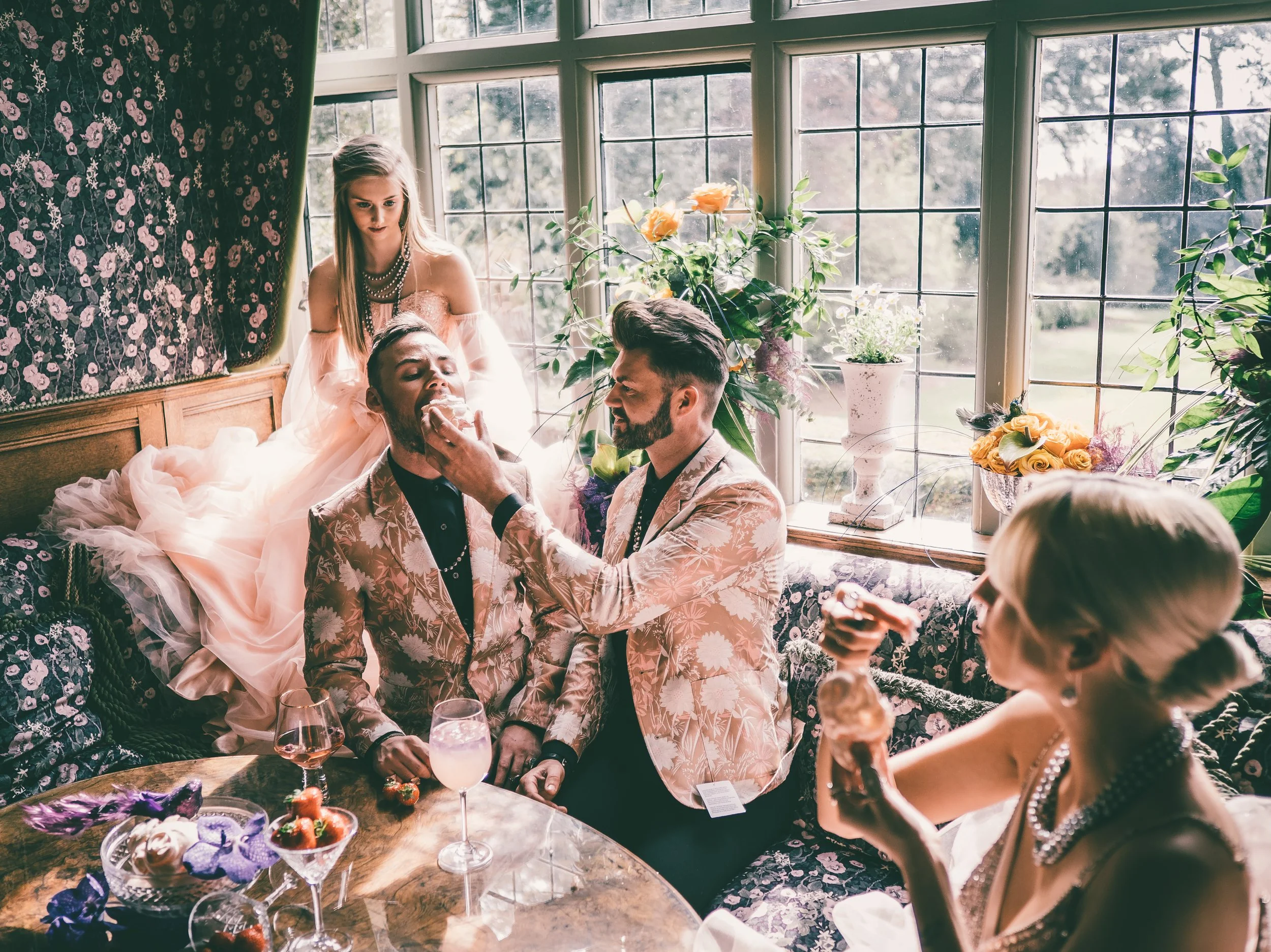 A group of people at a gathering, with one person feeding another, and others drinking, inside a room decorated with flowers and floral patterned furniture and curtains.