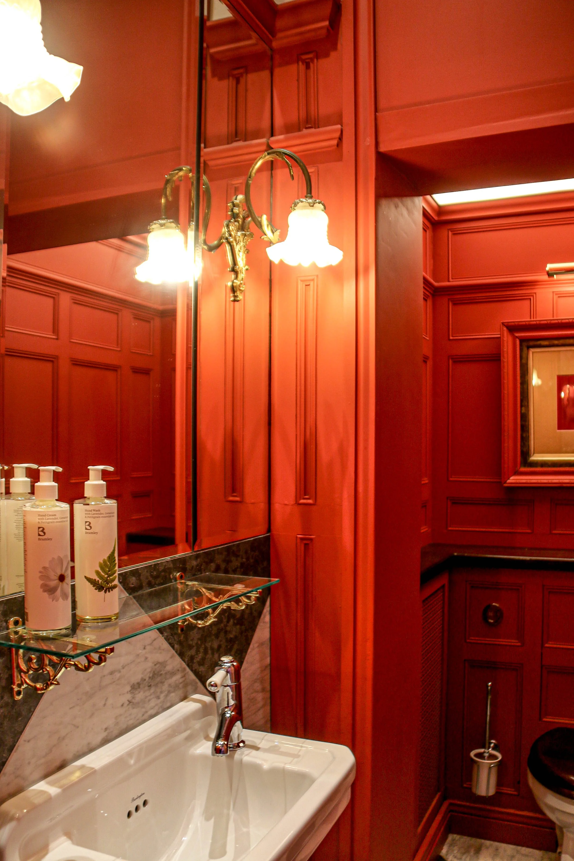 A small bathroom with red wooden-paneled walls, a white sink, a mirror, wall-mounted soap dispensers, and a decorative wall sconce with two lights.