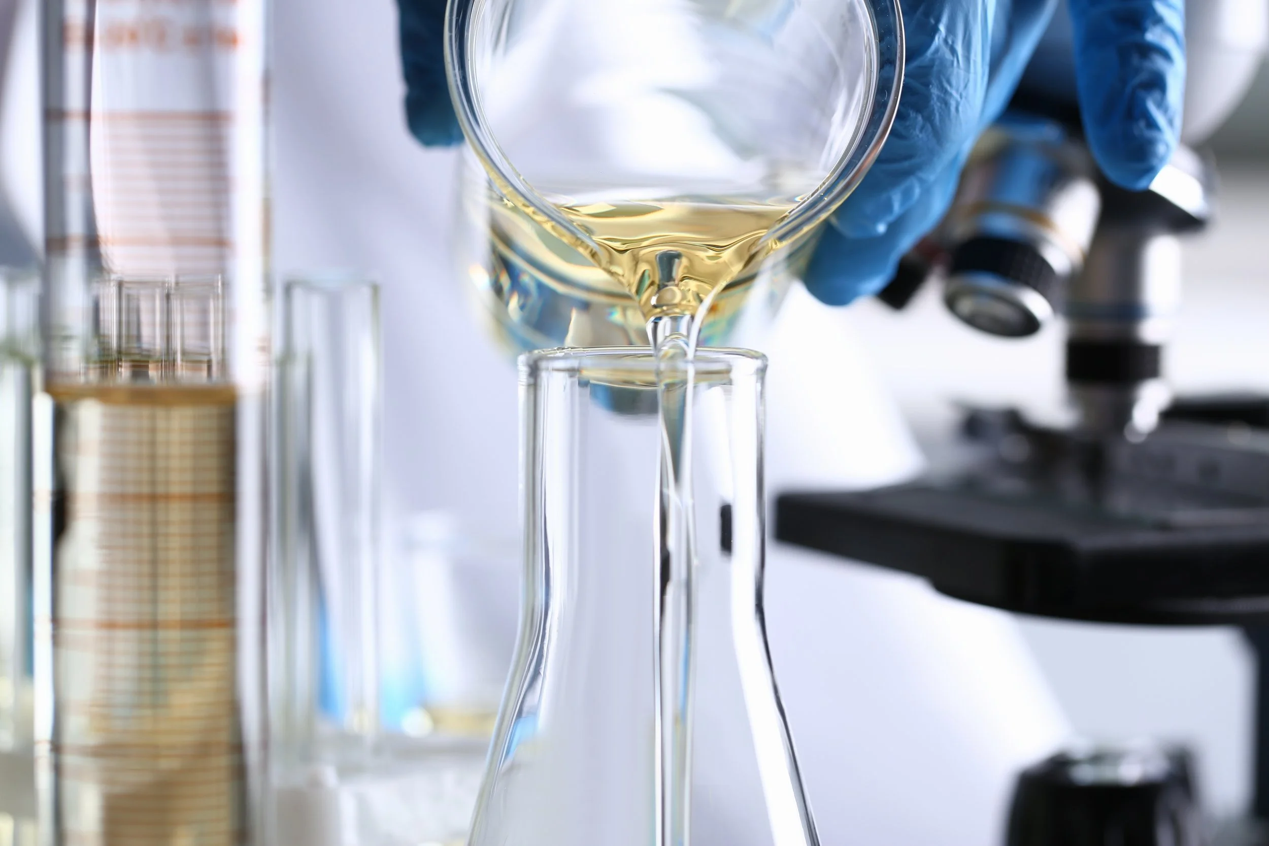 A scientist wearing blue gloves pours liquid into a glass flask in a laboratory setting with test tubes and a microscope in the background.