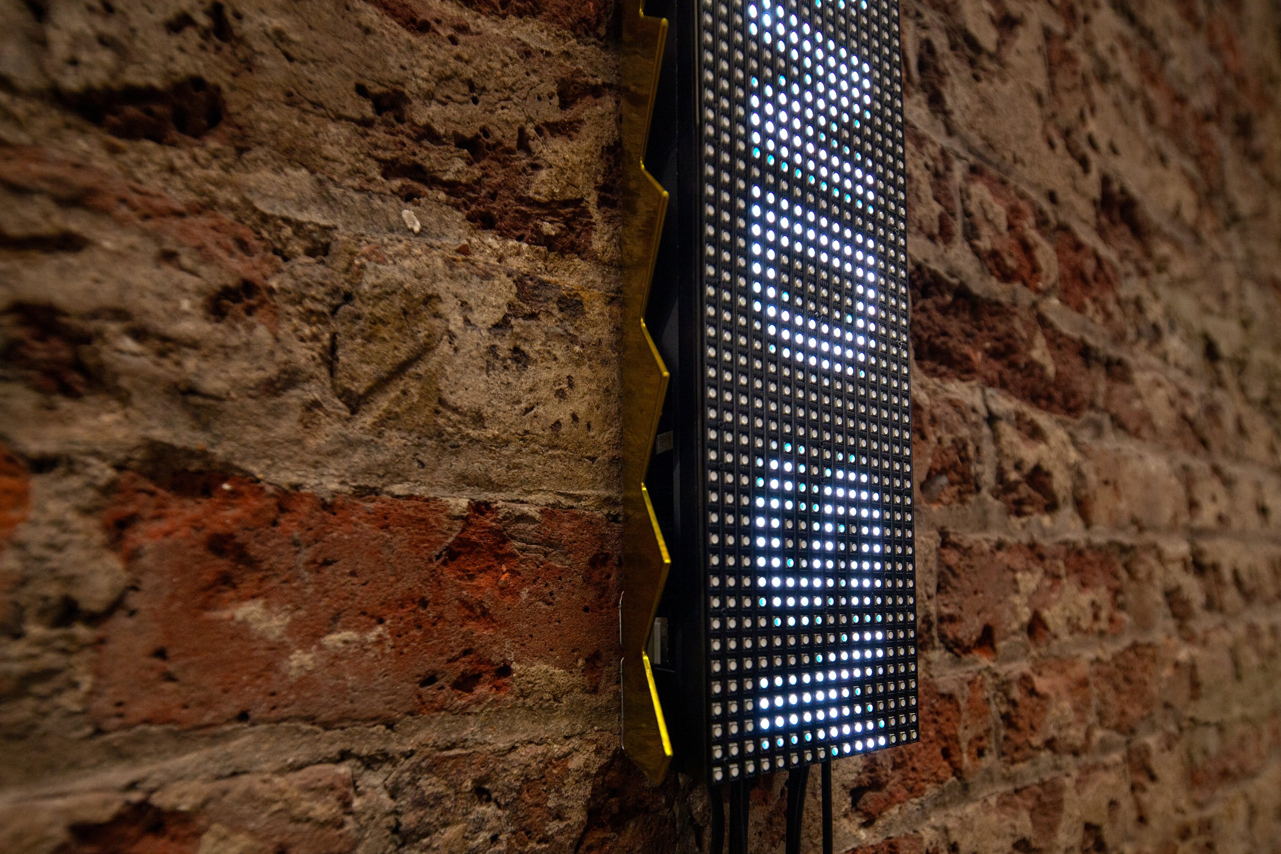 A narrow vertical LED ticker mounted on an exposed brick wall in a converted industrial interior, displaying rotated text, the display is encased in security spikes powder coated in gold